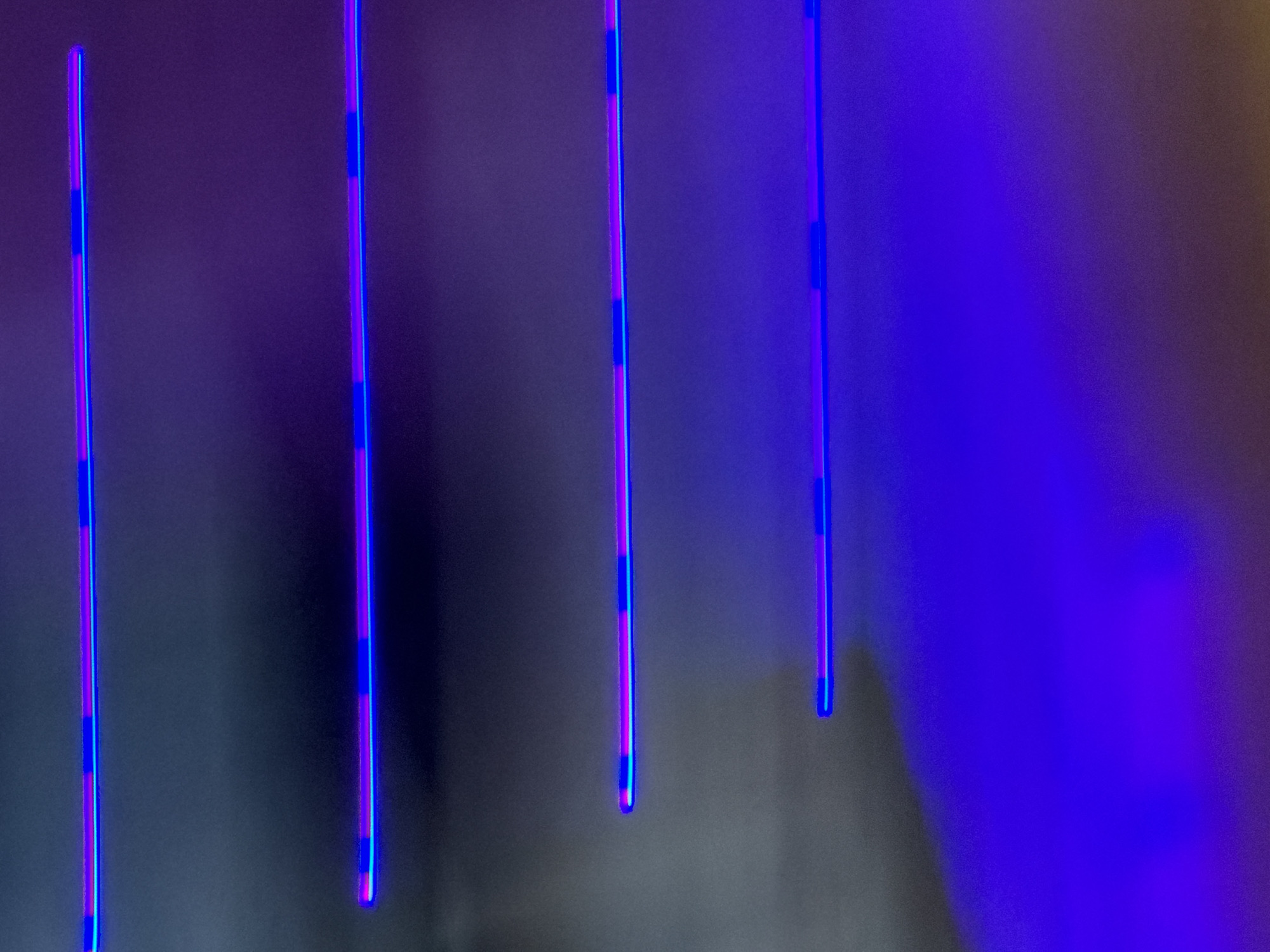 This is an abstract photograph featuring horizontal neon light strips against a dark, textured background. The light strips appear to be blue and pink, with segments of darker blue or purple appearing within the pink. The background transitions from a mottled grey on the left to a vibrant, luminous purple on the bottom right, suggesting an ambient light source. The image does not contain any recognizable landmarks or contextual clues that would place it in Bangkok, Thailand, or any specific city. The photograph is focused on the abstract play of light and color, with no people or discernible subjects present.
