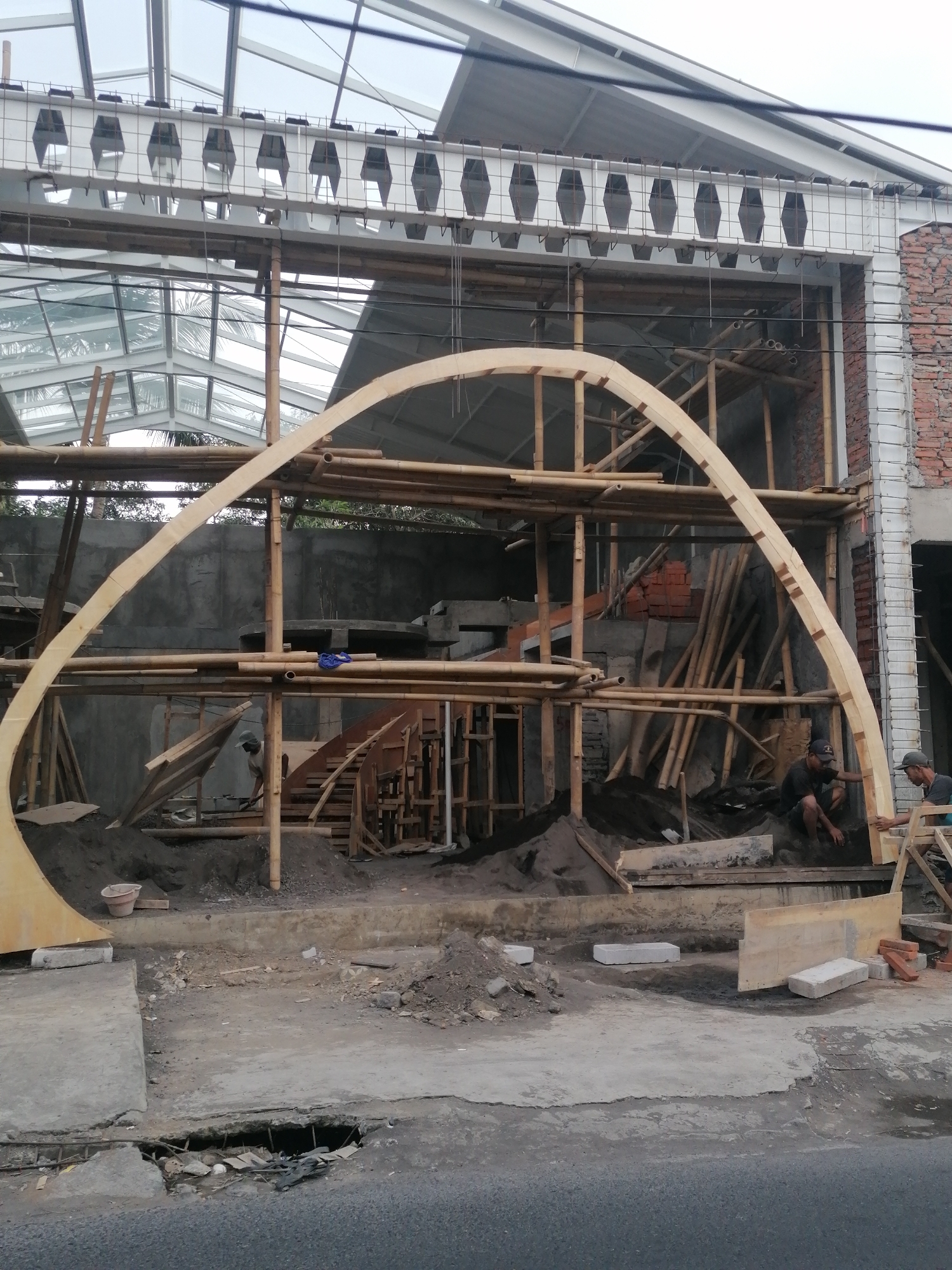 An unfinished building under construction in Ubud District, Indonesia, features a large curved wooden beam as a central structural element. The building's frame is composed of bamboo scaffolding and concrete. The surrounding area is a construction site with piles of sand, bricks, and building materials. Two men are visible, one appearing to be working with the wooden beam and the other nearby. A partially paved surface with a hole is in the foreground. The sky is overcast.