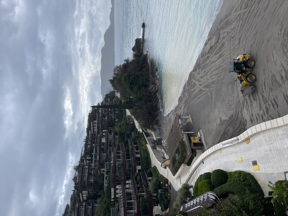 This image displays a coastal scene in Budva, Montenegro, under an overcast, grey sky. On the left and ascending a verdant hillside, a series of multi-story, terraced buildings with numerous balconies and integrated vegetation are visible. A curved, light-colored paved walkway with a low retaining wall winds along the base of these structures. Adjacent to the walkway and closer to the water, a structure with a thatched roof and another building are present on the land directly backing the beach. The foreground and mid-ground are dominated by a grey-sand beach, extensively marked by tire tracks. A yellow Caterpillar brand front-end loader is positioned on the beach, facing towards the lower left with its front bucket lowered. The light blue-green water of the sea occupies the right side of the frame, meeting the sandy shore and extending towards a rocky headland where a small, concrete pier or jetty protrudes into the water. Distant hills or mountains are discernible across the water under the cloudy conditions. No individuals are present in the scene.