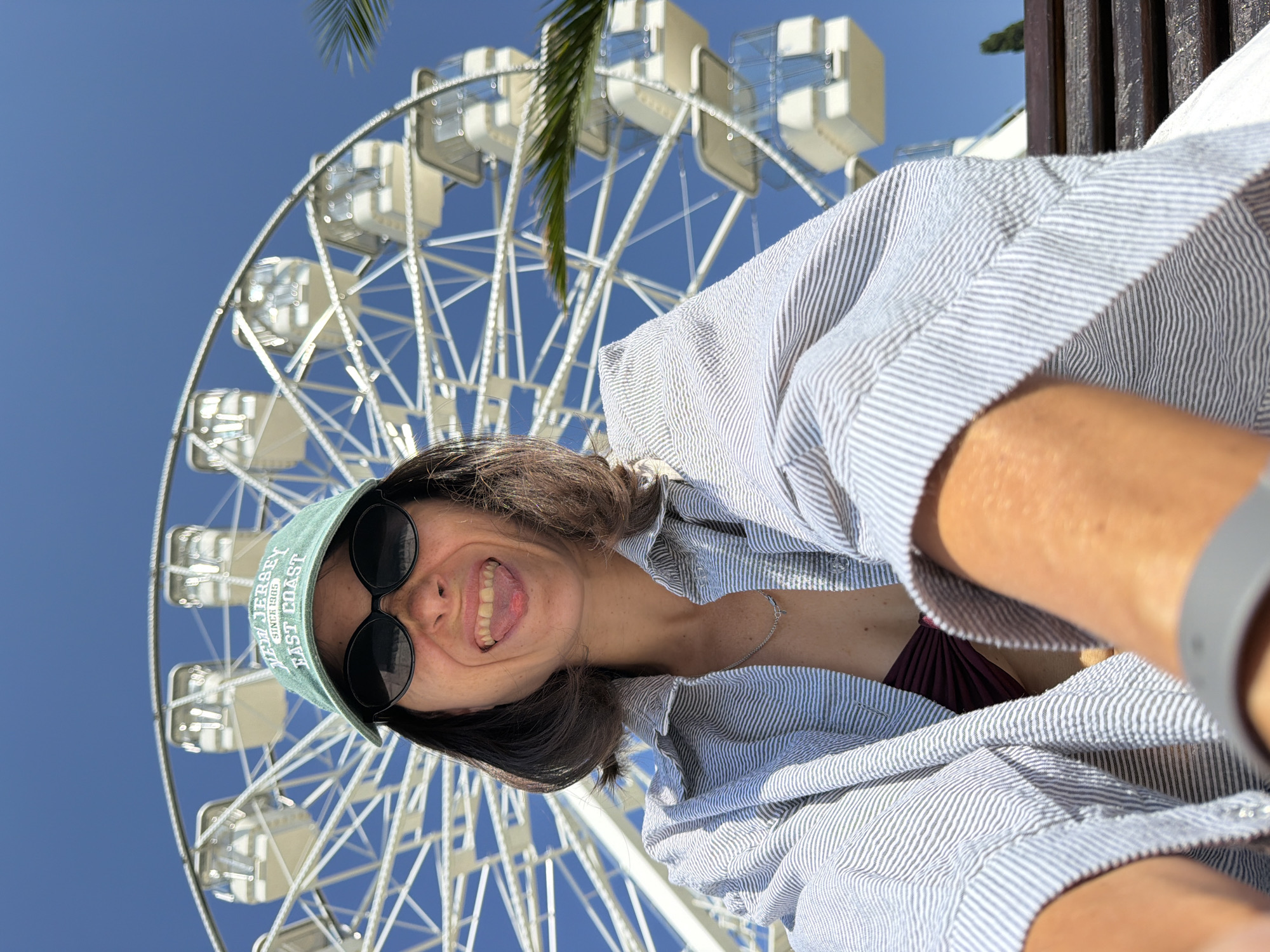 A woman is taking a selfie outdoors in front of a Ferris wheel. She is wearing a light blue and white striped button-down shirt over a burgundy bikini top, sunglasses, and a green baseball cap. The cap has text on it that reads "WEST JERSEY SINCE 1985 EAST COAST". She is smiling widely with her tongue sticking out, her mouth open, and her eyes wide. The Ferris wheel is white with many cabins and is set against a clear blue sky. A palm frond is visible in the upper left corner of the image. The sun is shining brightly, indicating it is daytime. The overall mood of the image is cheerful and playful.