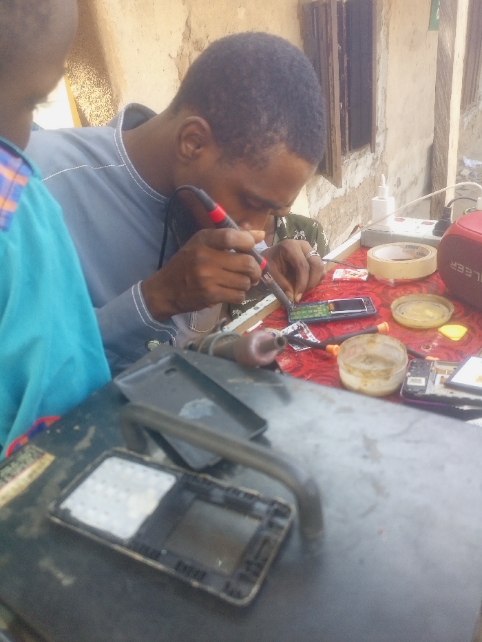 An outdoor mobile phone repair scene in Akwanga, Nigeria, depicts an adult male intently soldering an electronic component on a small circuit board, likely from a mobile phone. He wears a grey long-sleeved shirt and holds a red-and-black soldering iron in his right hand, with his left hand steadying the device. Partially visible to his left, another individual in a blue shirt observes the process. The workspace features a table covered with a red patterned cloth, laden with various electronic repair tools and components. These include a disassembled feature phone, additional phone parts, small screwdrivers, two clear containers holding yellowish soldering flux, a roll of adhesive tape, a white power strip connected to a red "GLEER" brand portable speaker, and several small wires and circuit boards. In the immediate foreground, a dark surface holds an open mobile phone frame with internal components exposed and a dark phone case. The background consists of a weathered, light-brown plastered wall with a small, dark rectangular window opening, indicating an informal outdoor or semi-outdoor setting.