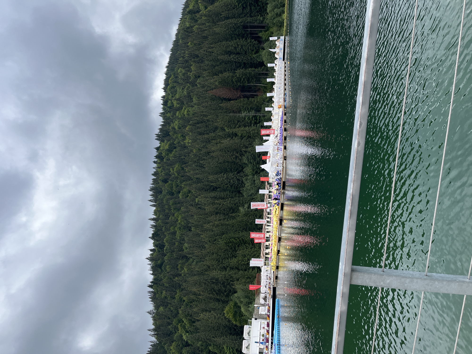 The image depicts a serene scene at Bukovel resort in Polianytsia, Ukraine. The foreground is dominated by a dark green body of water, likely a lake, with ripples reflecting the overcast sky. A metallic railing with wire ropes stretches across the bottom of the frame, suggesting a viewing platform or walkway.
In the middle ground, a wooden dock extends into the water, lined with white tents and canopies. Numerous red and white flags with text are planted along the dock, indicating a festive or event setting. Yellow and purple inflatable structures are arranged on the water near the dock. Several small, colorful boats are visible on the water.
The background is a dense, green coniferous forest covering a steep hillside. The sky is heavily overcast with dramatic, dark clouds, suggesting impending rain or a storm. The overall atmosphere is one of natural beauty enhanced by a man-made event setup.
