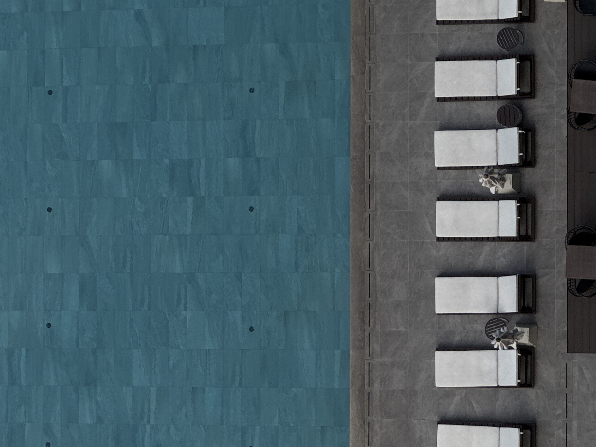 This is an aerial, top-down view of a resort swimming pool and its surrounding deck, likely located in Bangkok, Thailand. The pool itself is large, rectangular, and filled with clear turquoise water, tiled with a pattern of dark teal squares. Several circular drains are visible on the pool floor.
Adjacent to the pool is a wide, grey tiled deck area. Along the edge of the pool, there is a grey stone border with a series of dark, rectangular drainage grates.
On the deck, there are seven white lounge chairs with dark wooden frames, positioned in a horizontal row. Most of these chairs are empty. One chair, second from the left, has a small decorative sculpture of a flower on a pedestal next to it, along with a small, dark circular side table. Another chair, fourth from the left, also has a similar small, dark circular side table next to it.
Further down from the lounge chairs, a dark wooden deck area is visible, with what appear to be dark wicker chairs and tables, though these are partially cut off from the frame.
The overall scene is serene and unoccupied, suggesting a tranquil resort environment.