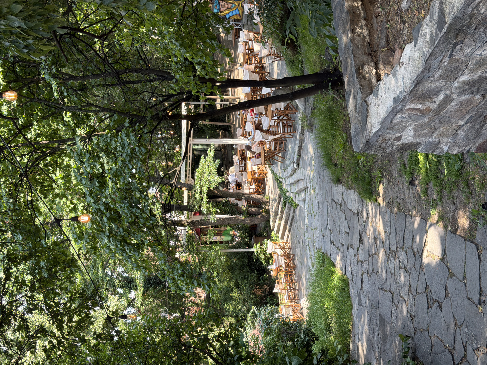 The image displays an outdoor cafe or restaurant situated within a heavily treed, garden-like environment, characteristic of urban green spaces in Plovdiv, Bulgaria. A winding pathway, paved with irregularly shaped dark grey flagstones, extends from the foreground towards the midground, receiving dappled sunlight. To the right of the pathway in the foreground, a low stone wall defines a raised planting bed. The main dining area occupies the midground and background, featuring numerous square wooden tables paired with folding chairs that have white canvas seats and backs. These tables are arranged on various levels, with stone steps providing access to elevated sections. Several patrons are visible seated at these tables, engaged in dining and conversation. Overhead, multiple strands of decorative string lights, fitted with exposed warm-toned Edison-style bulbs, are suspended between the prominent tree branches. The scene is densely canopied by mature trees with lush green foliage. In the upper right background, a portion of a colorful mural depicting a stylized figure with a blue headdress is visible on a vertical surface.