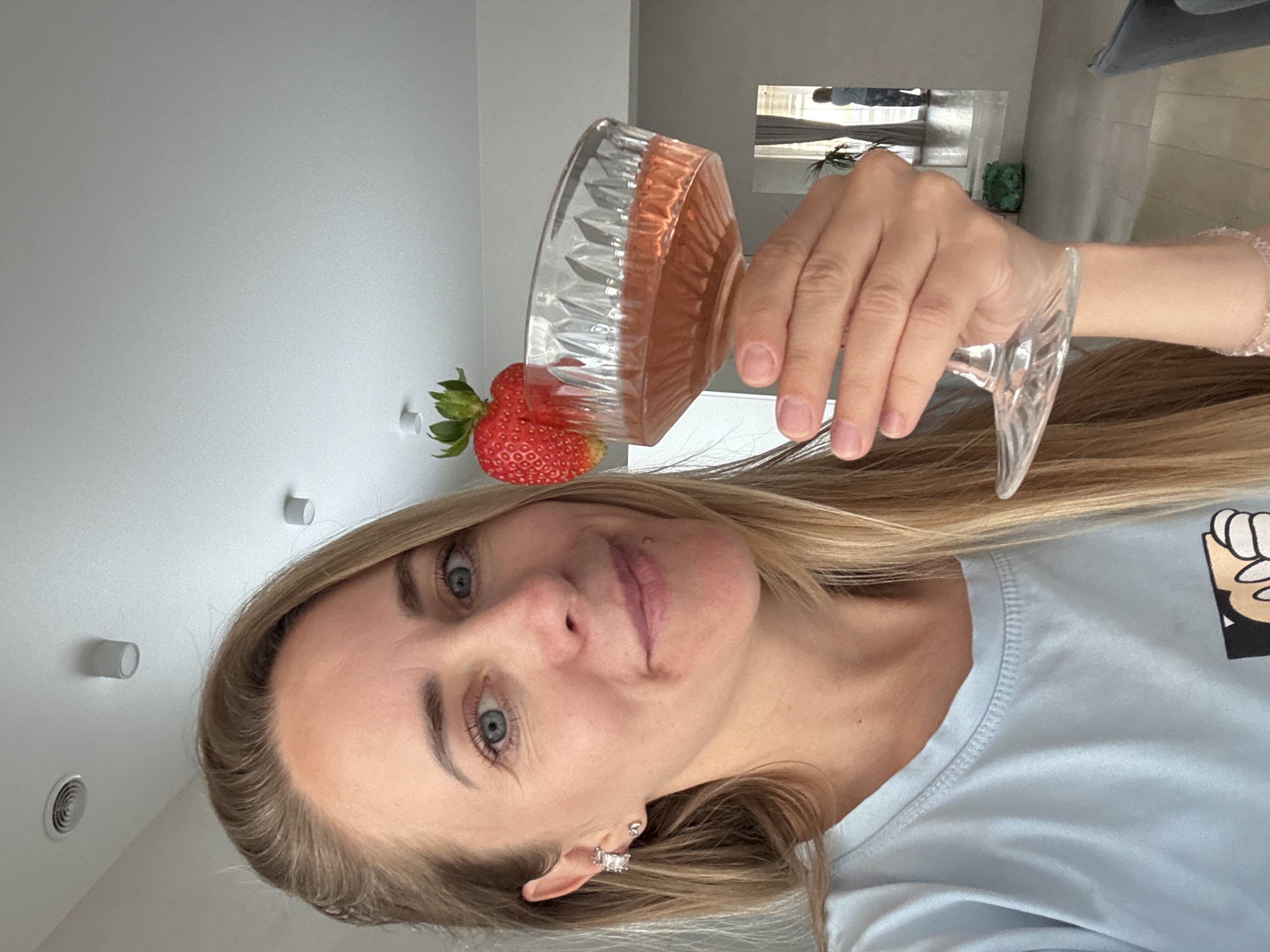A woman with blonde hair is holding a glass of rose wine, garnished with a strawberry. She is wearing a light blue t-shirt and a sparkly bracelet on her wrist. The background shows a room with white walls and a light-colored floor. In the distance, a person is visible through a doorway. The image appears to be taken indoors in a residential setting. There is no indication of the specific location within the city of Chaikovsky, Russia.