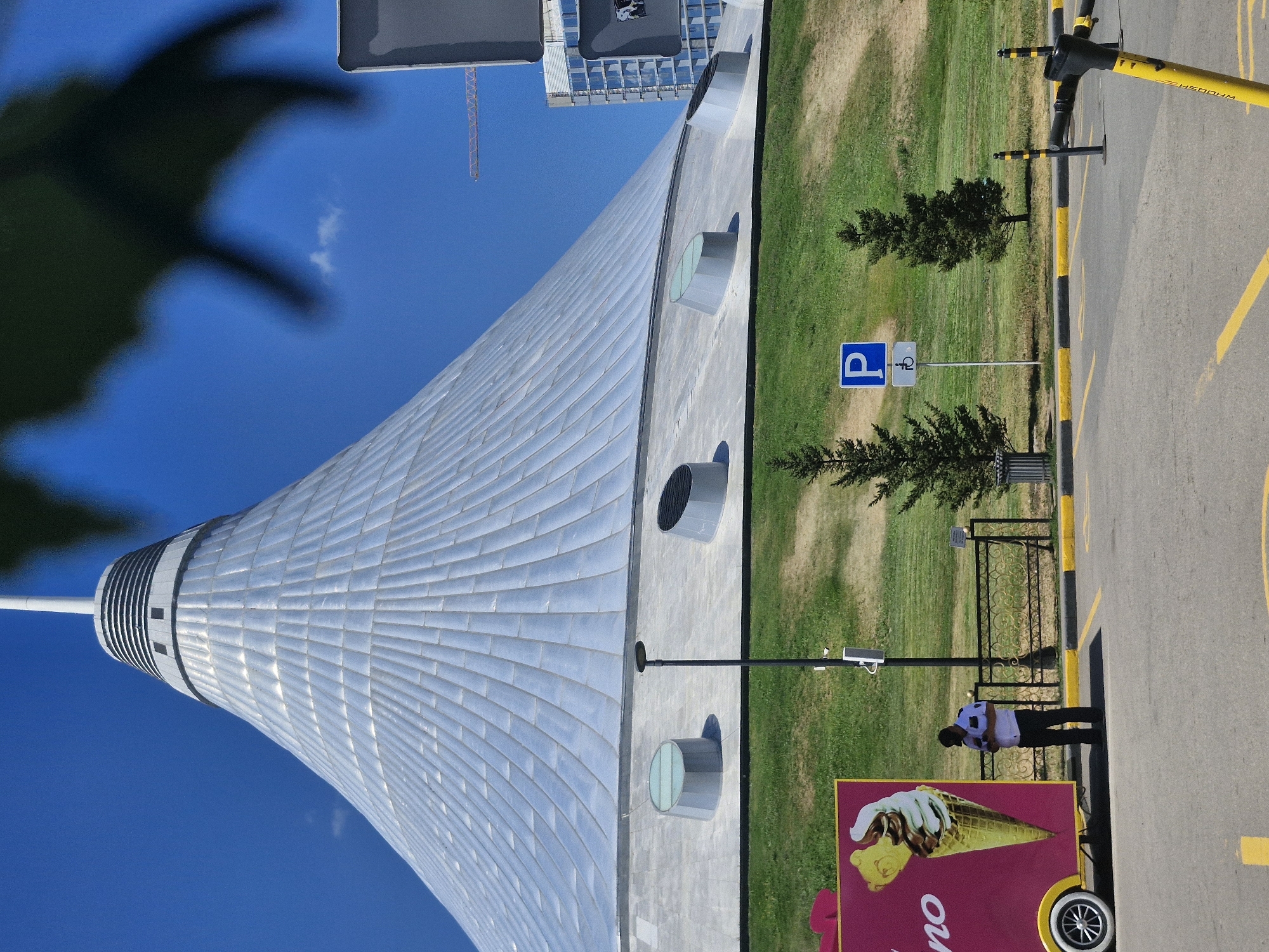 The image depicts the Khan Shatyr Entertainment Center in Astana, Kazakhstan, on a clear, sunny day. The iconic tent-like structure, with its metallic, ribbed facade, dominates the upper portion of the frame. Below the structure, a green, sloping lawn is visible, interspersed with a few evergreen trees. In the foreground, a paved area, likely a parking lot, is marked with yellow lines.
On the left side of the image, a bright pink ice cream cart is parked, featuring a large graphic of a swirled ice cream cone. A man, dressed in a white short-sleeved shirt, dark pants, and a black cap, stands near the cart, appearing to be looking at his phone. To the right of the man, a lamppost stands tall. Further to the right, a blue parking sign with a white 'P' and a blue sign with a white wheelchair symbol are mounted on a pole. Two small, conical evergreen trees flank this sign.
In the background, a modern glass-fronted building and a construction crane are visible, suggesting ongoing development in the city. A dark, leafy branch is blurred in the upper right corner, partially obscuring the sky and the top of the tent. A yellow electric scooter is parked on the right side of the paved area, its handlebars prominent in the foreground. The overall scene is one of urban activity set against a backdrop of distinctive architecture.