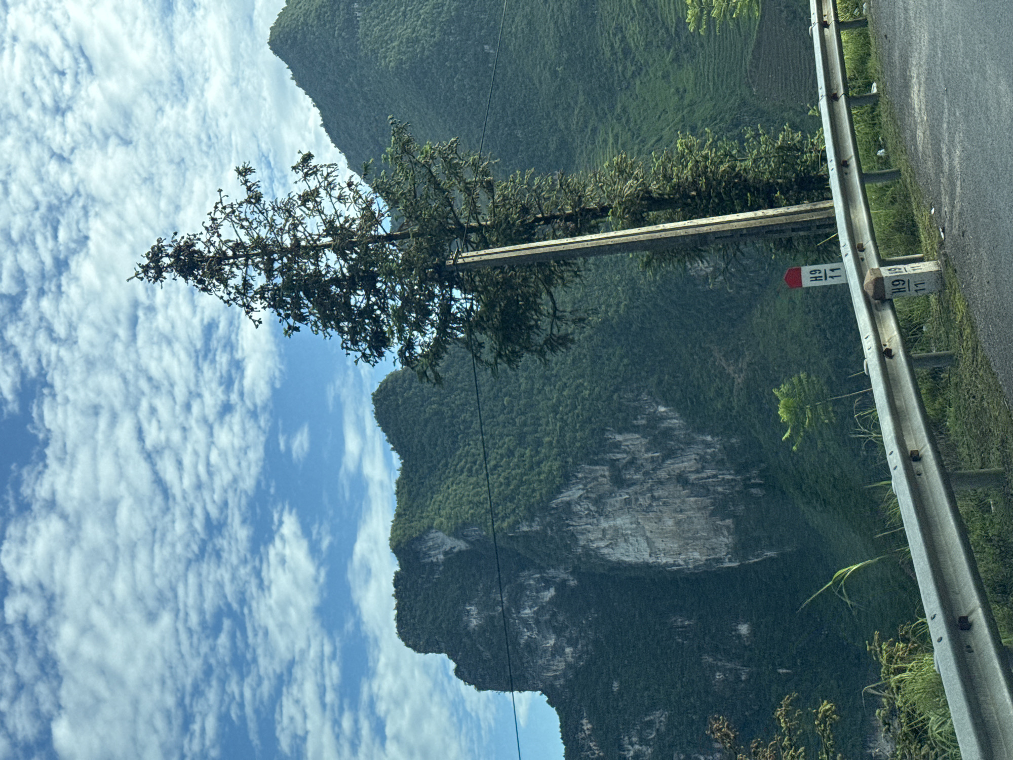The scene is a roadside view in Lũng Cú Commune, Vietnam. A guardrail runs along the right side of the image next to an asphalt road. Roadside markers are placed next to the guardrail. A power pole stretches across the frame, with power lines extending out of the frame. A tree is growing next to the pole. The background features a lush, green, mountainous terrain. Above the mountains is a partly cloudy blue sky. The image is taken from a low angle and rotated to the right.