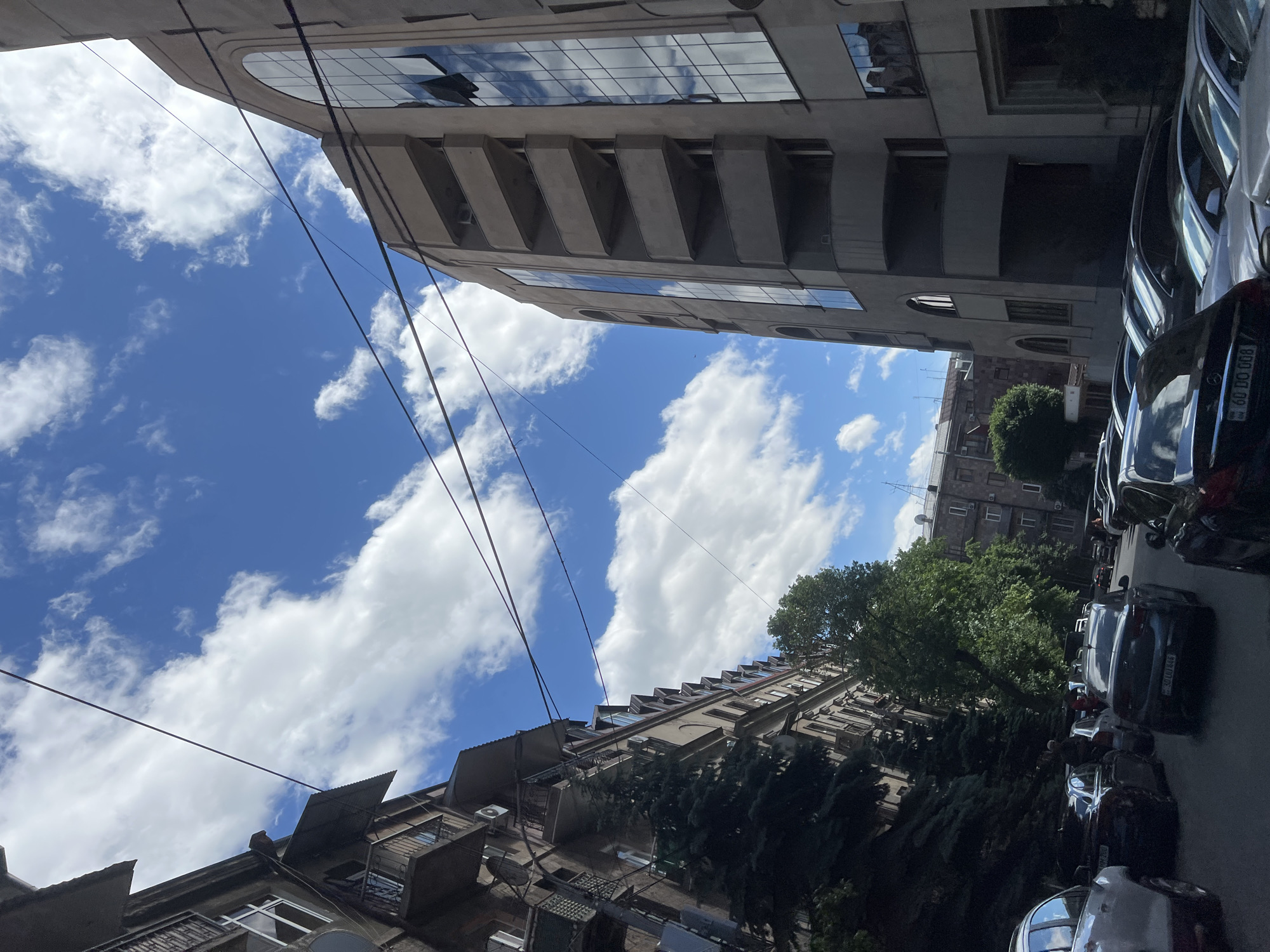 The image depicts an urban street scene in Yerevan, Armenia, viewed from a low angle looking upwards. Two tall buildings flank the sky. The building on the left is older with protruding architectural elements like balconies and air conditioning units. The building on the right appears more modern, featuring reflective glass panels and a stepped façade. A row of parked cars is visible on the right side of the street. Trees with green foliage intermix with the buildings. Power lines crisscross the open sky, which is blue with scattered cumulus clouds. The overall impression is a typical urban environment.