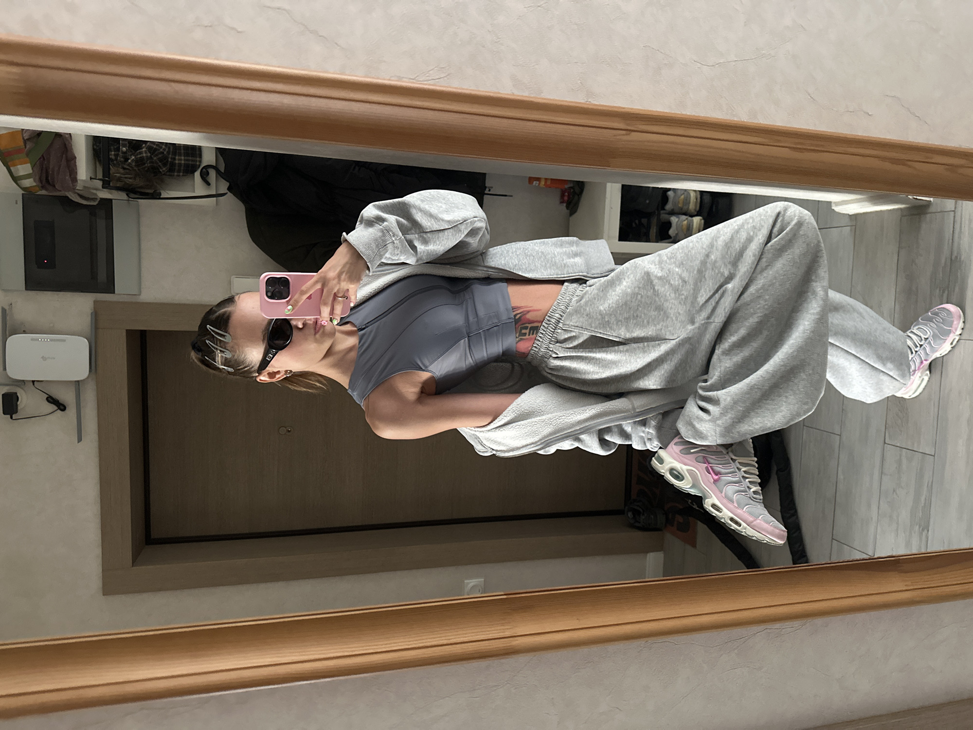 A woman is taking a mirror selfie in a hallway with a wooden-framed mirror. She is wearing a grey sports bra, a grey zip-up hoodie, and grey sweatpants. Her hair is pulled back with silver clips, and she is wearing black sunglasses and a pink phone case. Her left leg is bent and raised. The hallway appears to be in an apartment, with a door visible behind her and shelves with items on the right.