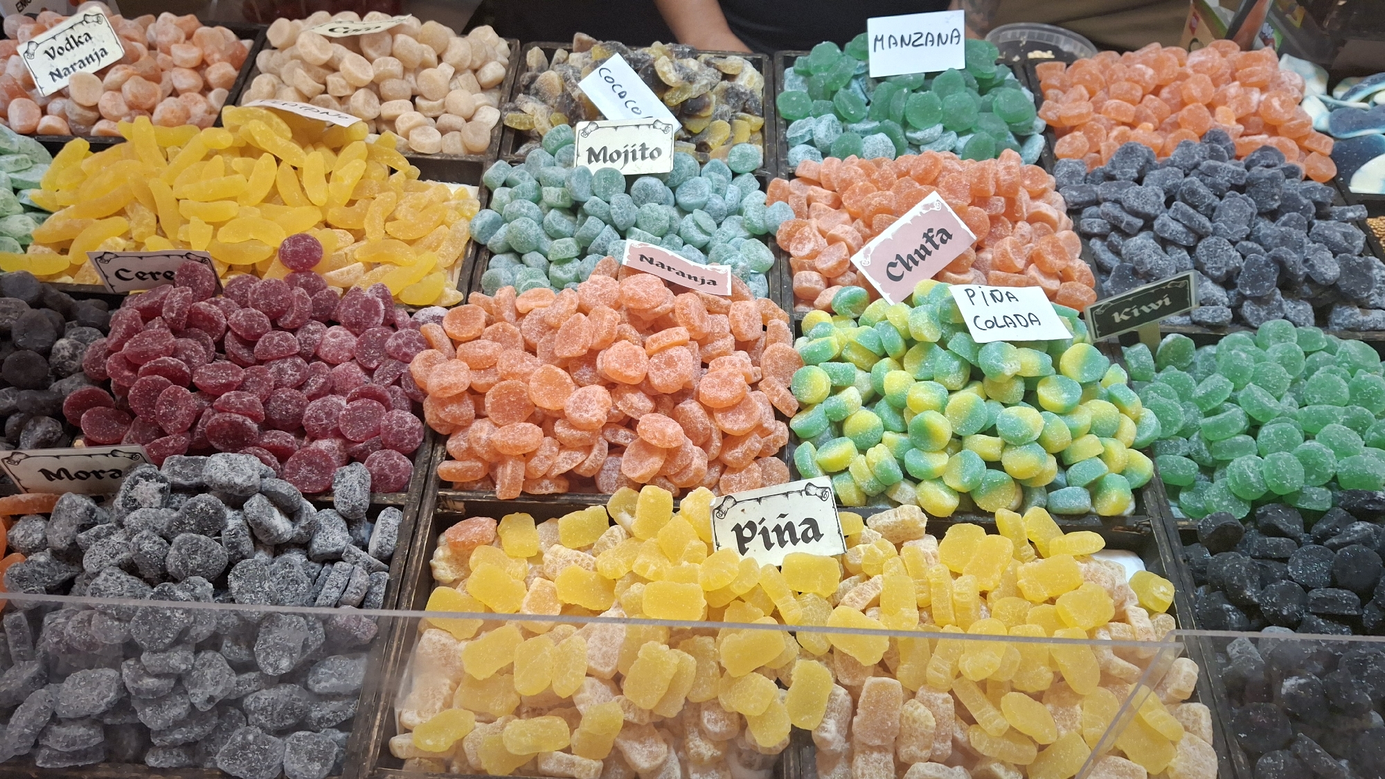 The image displays a vibrant market stall in La Fresneda / la Freixneda, Spain, filled with a colorful assortment of gummy candies. The candies are arranged in individual wooden crates, each labeled with its flavor. Visible labels include "Vodka Naranja", "Cere", "Mora", "Mojito", "Manzana", "Naranja", "Chufa", "Piña Colada", and "Kiwi". The candies themselves vary in shape and color, with prominent displays of orange, yellow, red, green, blue, and black varieties. The lighting suggests an indoor or shaded outdoor market setting. No people are clearly visible in the frame, and the focus is entirely on the confectionery display. The scene is presented in a close-up view, highlighting the textures and details of the candies.