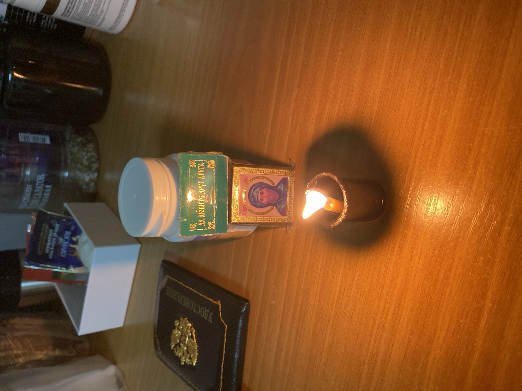 An overhead, slightly angled shot captures a collection of objects arranged on a warm-toned wooden surface, illuminated by the glow of a burning candle. In the foreground, a dark blue or black book with gold trim lies flat, featuring a golden embossed emblem of a double-headed eagle on its cover. Cyrillic text, "УДОСТОВЕРЕНИЕ" (Udostovereniye, meaning "Certificate" or "Identification"), is visible along its spine.
To the left of the book, a small white open box contains a blue packet with the brand name "RICHARD" and "ROYAL ENGLISH STRAWBERRY" printed on it. Further back, a white plastic bottle with a white screw-top lid stands upright. A small, green matchbox-like item, bearing Cyrillic text "ДА ЛЮБИТЕ ДРУГ ДРУГА" (Da Lyubite Drug Druga, meaning "Love Each Other") and "2023", is placed next to the bottle. Attached to this green box, or resting near it, is a small, golden-framed icon depicting a religious figure, likely the Virgin Mary or a saint, with a serene expression.
A lit candle in a dark holder stands to the right of the icon, its flame casting a warm, flickering light that creates highlights and shadows across the wooden surface. In the blurred background, several dark glass jars or bottles are visible, out of focus.
The scene suggests a quiet, possibly personal or devotional setting, with the candle as a focal point, likely indicating prayer, remembrance, or simply providing ambient light. The presence of the icon and Cyrillic texts points to an Orthodox Christian or Eastern European cultural context. The time of day appears to be evening or night due to the prominent use of candlelight.