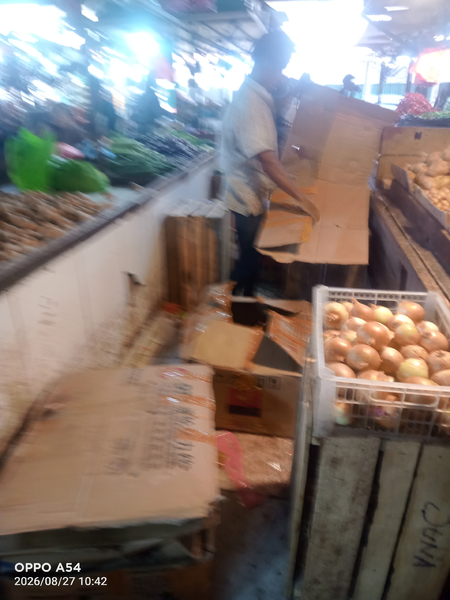 The image depicts a market scene, likely in Jambi, Indonesia, with a focus on the display and handling of produce. In the foreground, a white plastic crate is filled with onions. To the left, several cardboard boxes are stacked, some appearing to contain goods. A person, partially visible, is in the process of handling a large cardboard box. In the background, a variety of fresh produce, possibly vegetables like leafy greens and root vegetables, are arranged on stalls. The overall environment suggests a bustling marketplace with goods being moved and organized. The lighting indicates it is daytime, possibly indoors or under a covered area typical of markets.
