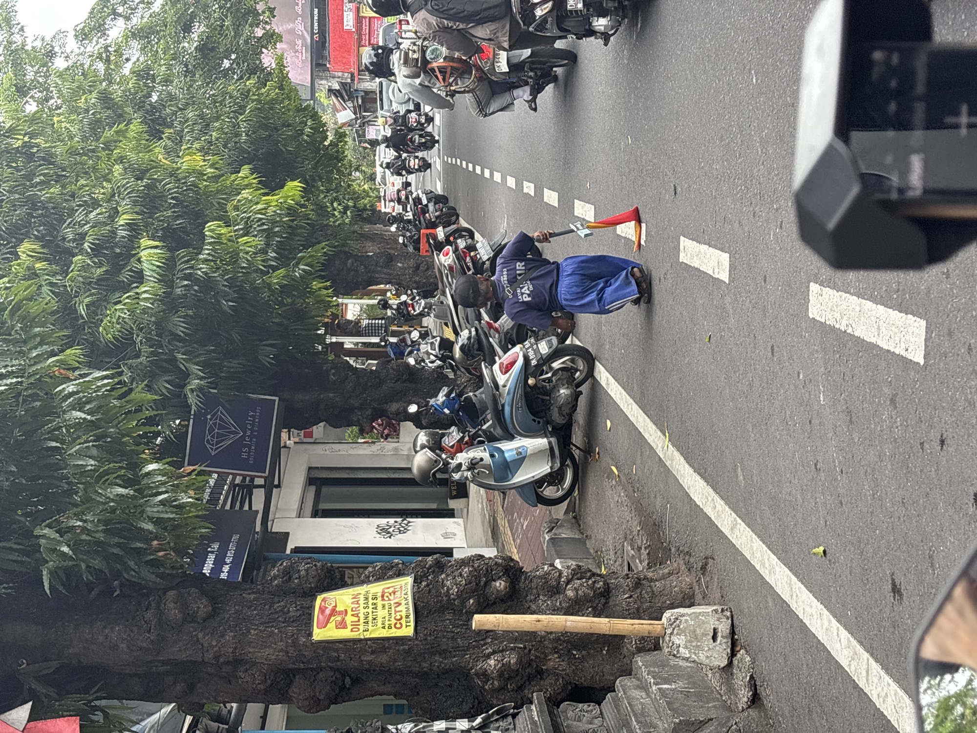 The image captures a street scene in Denpasar, Indonesia, likely a busy commercial area given the presence of shops and numerous motorcycles. The foreground shows a person of short stature walking away from the camera down the right side of the road, wearing a dark blue t-shirt with white lettering and blue pants. They are holding a tool that appears to be a broom or mop with a red flag attached to the end. The road is paved and marked with white lines, indicating lanes and pedestrian crossings.
Along the left side of the street, there are large trees with substantial trunks and lush green foliage, casting shadows on the sidewalk. A yellow sign with red lettering is attached to one of the trees, with text that appears to be a warning or directive. Several parked motorcycles are visible along the sidewalk, and a blue and white scooter is prominent near the center left.
In the background, more motorcycles are in motion, some carrying riders with helmets. Buildings line the street, with shop signs visible, including one for "HS Jewelry Goldsmiths & Diamonds." The overall atmosphere suggests a typical day in a bustling urban environment. The perspective is from a slightly elevated viewpoint, possibly from another vehicle or motorcycle.