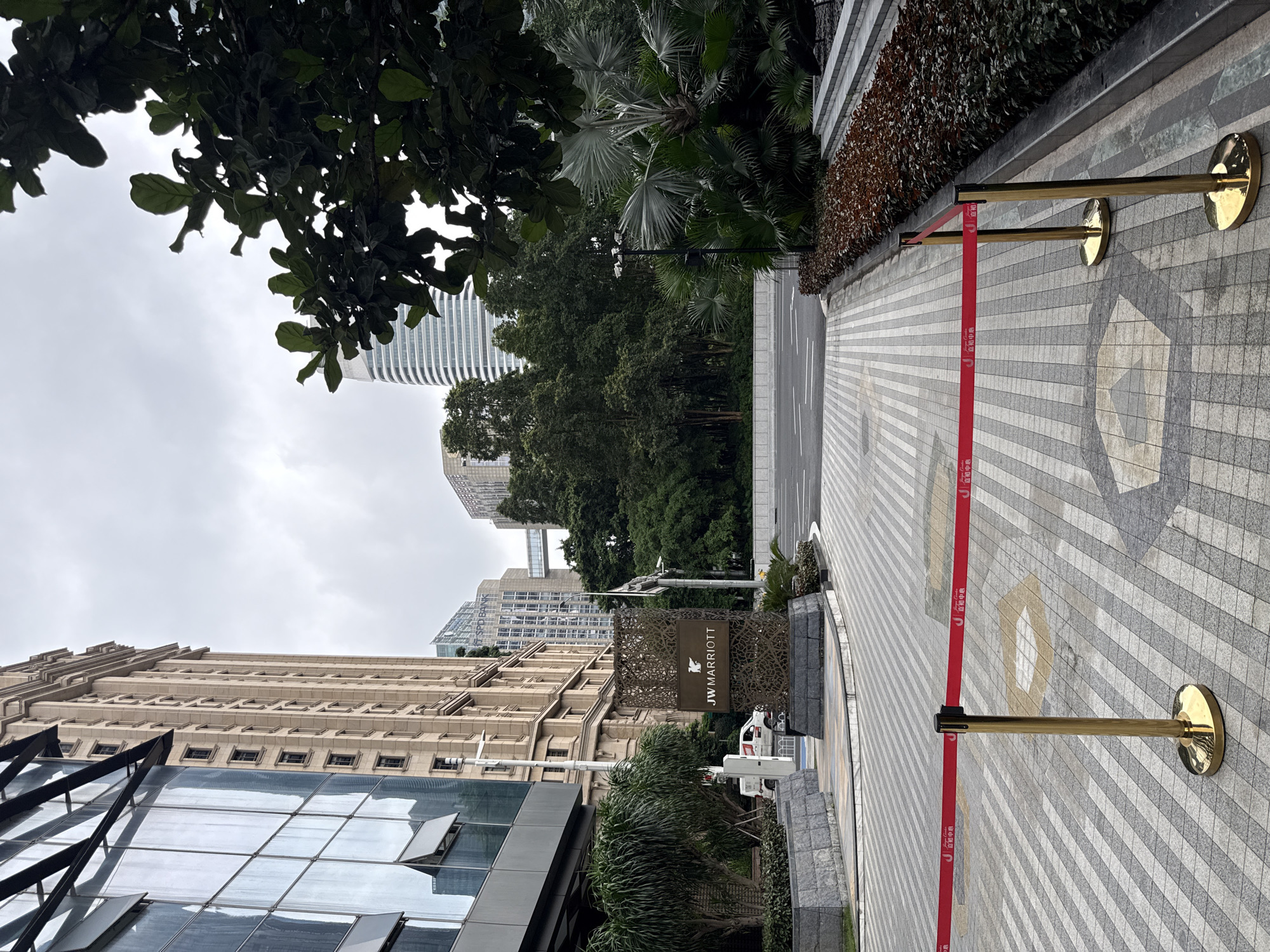This image captures an outdoor scene in Tianhe District, China, featuring the entrance to the JW Marriott hotel. The foreground is dominated by a patterned tiled walkway, bisected by a red velvet rope barrier supported by gold stanchions. To the left, the modern facade of a building with large glass windows and a darker, angled structure is visible, reflecting the overcast sky and surrounding architecture. Adjacent to this building is the JW Marriott hotel's signage, a decorative metal structure with the hotel's logo and name. Behind the signage, a white utility vehicle is parked.
The midground shows a lush green environment with various trees and palm-like plants, suggesting a well-maintained landscape. A road with white lane markings is visible, separating the hotel entrance area from further structures. In the background, a tall, modern skyscraper with a curved, white facade stands out against the cloudy sky. Other buildings, including one with the "SPD BANK" logo, are also present, indicating a commercial or business district. The overall atmosphere is subdued, likely due to the cloudy weather. No people are visible in the frame.