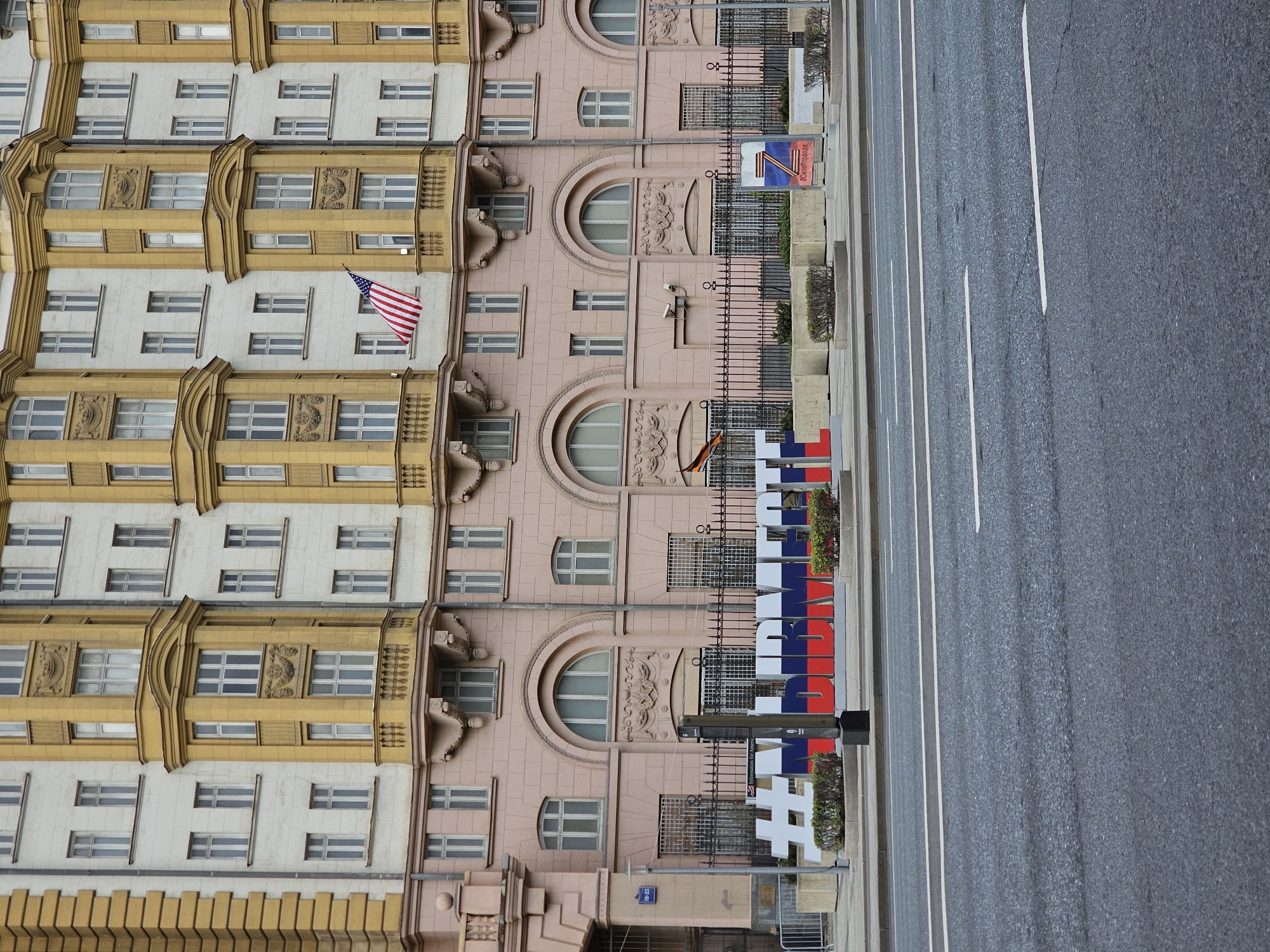 The image depicts a street scene in Moscow, Russia. A multi-story building is seen to the left, with varying architectural styles on different sections, including yellow and pink facades, and numerous windows. An American flag hangs from one of the windows. Along the sidewalk, near the building, there are large, white block letters spelling "#VISITMOSCOW" with red, white, and blue coloring. Adjacent to the sidewalk is a multi-lane road. The overall environment appears urban.