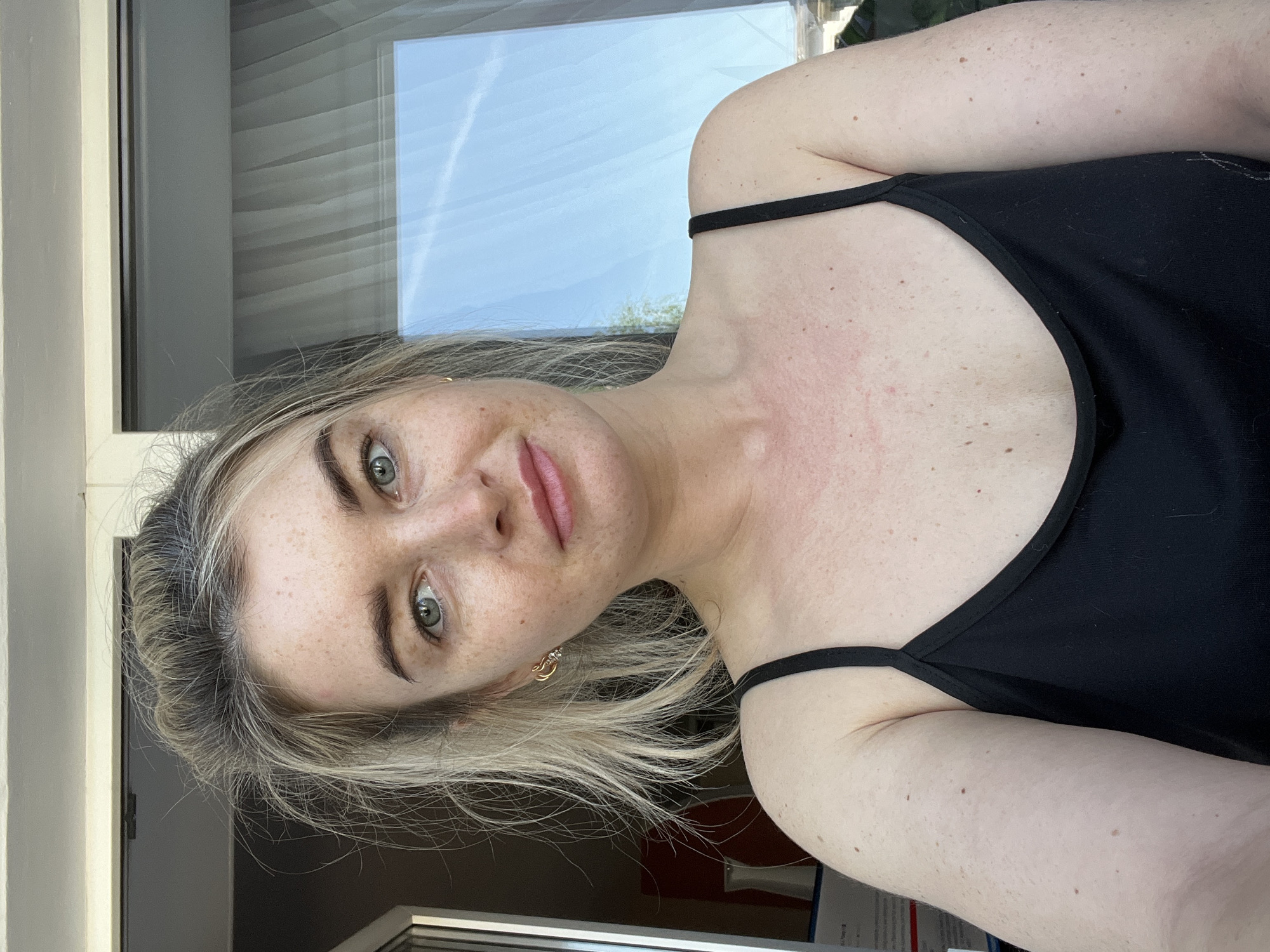 A fair-skinned woman with blonde hair and freckles is the subject of this portrait-style photograph. She is wearing a black tank top with thin straps. The woman's face is directed towards the camera, and she has a neutral expression.
Behind the woman, there is a window. The reflection in the window suggests a bright sky with some cloud cover.
It is impossible to ascertain the location of this image as being in the city of Chaikovsky, Russia, with the given information. There are no elements in the image that definitively identify it as being in Chaikovsky, Russia.