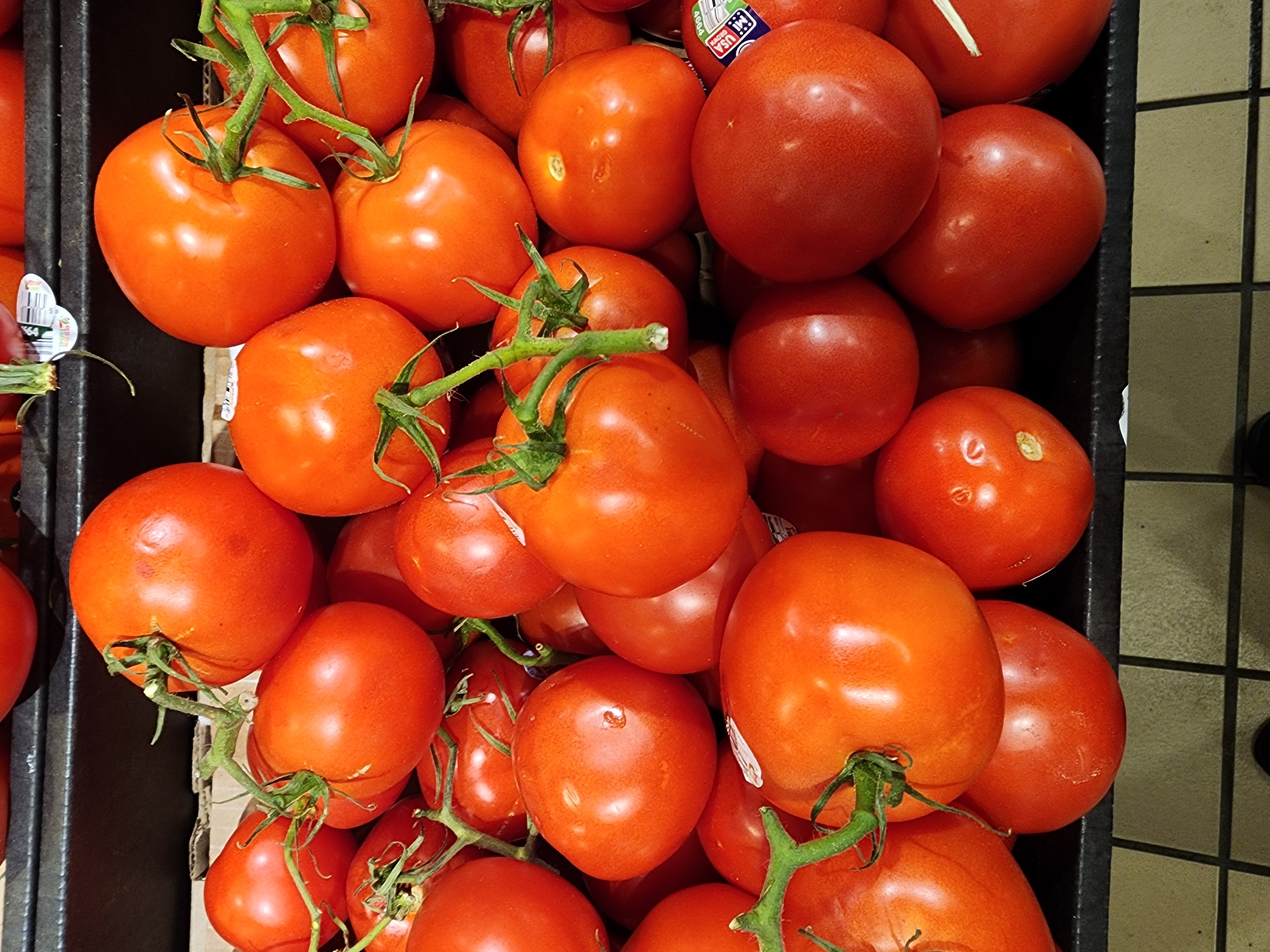 A close-up, eye-level, overhead shot displays a large pile of ripe, red tomatoes arranged in a retail display. The tomatoes are predominantly round and smooth, with some showing slight imperfections like dents or blemishes. Several tomatoes have green stems attached, some still clustered together, indicating they are vine-ripened. The lighting highlights the glossy sheen of the tomato skins, with some appearing more vibrantly red than others. The tomatoes are packed tightly together, filling the frame from edge to edge, with the bottom of the image showing a dark, textured surface that appears to be a shelf or bin, possibly with a grid pattern. The background is dark and out of focus, suggesting a commercial setting like a grocery store.