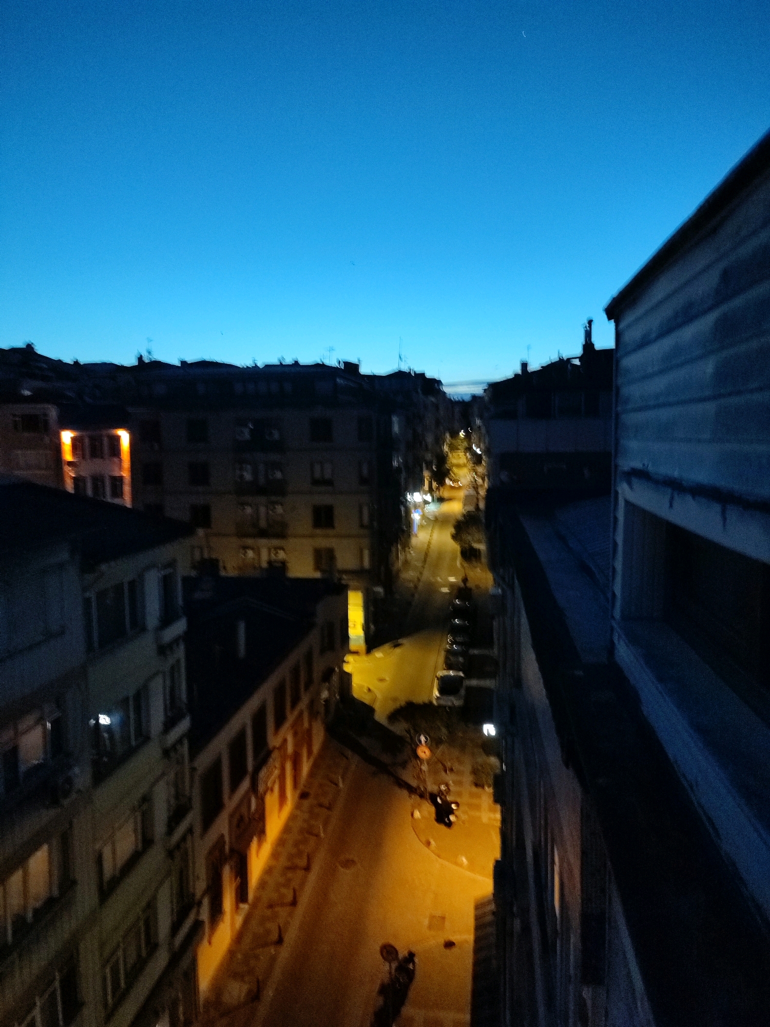 The image captures a street scene at dusk or dawn in Kadıköy, Turkey. The sky is a deep, clear blue, transitioning to lighter shades near the horizon. On the right, a building with a textured, possibly wooden or siding facade dominates the foreground, with a visible window. To the left, a street lined with multi-story buildings recedes into the distance. Streetlights cast a warm yellow glow on the asphalt road and illuminate the facades of the buildings, some of which have visible windows. Several vehicles, including cars and what appear to be motorcycles, are parked or in motion on the street. The architecture suggests an urban environment, with the buildings close together. The overall impression is of a quiet city street transitioning from daylight to evening, or vice versa, with signs of human activity from the illuminated windows and vehicles.