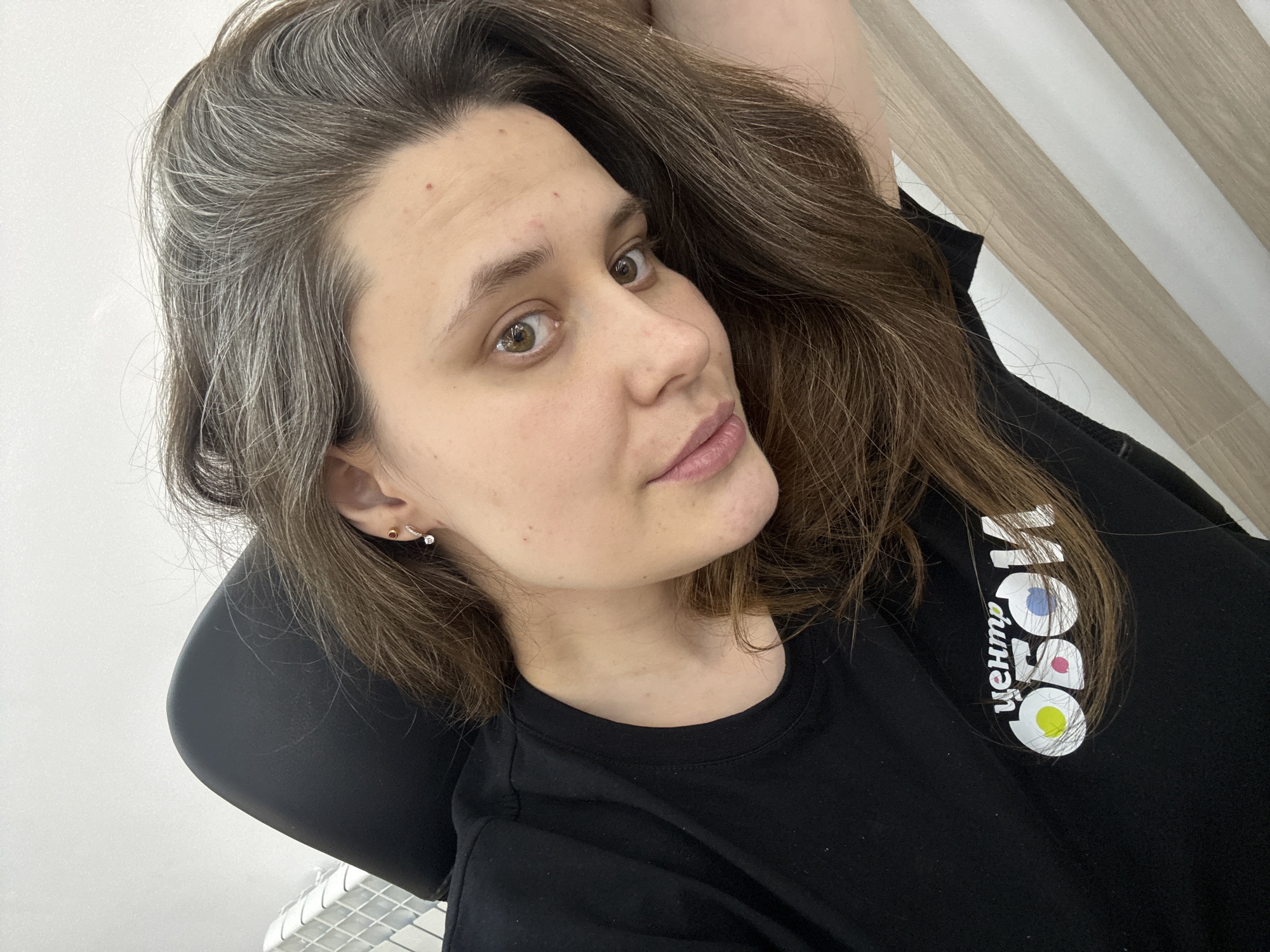 A medium-shot selfie of a woman with light skin and hazel eyes. Her hair is shoulder-length, with grey at the roots and brown at the ends. She wears two earrings in her left ear. She is wearing a black t-shirt with a white design and the Cyrillic word "Центр" (Tsentr) on it. The design includes a blue, a pink, and a yellow circle. The background shows what appears to be a wall of light wood paneling. A portion of what looks like a black chair is in the lower-left corner.
The brand on the t-shirt, "Центр Мой Документы" or "My Documents Center" is a Russian government service. These centers are located in most cities in Russia, including the city of Chaikovsky. It is therefore very likely the photograph was taken in Chaikovsky, Russia.