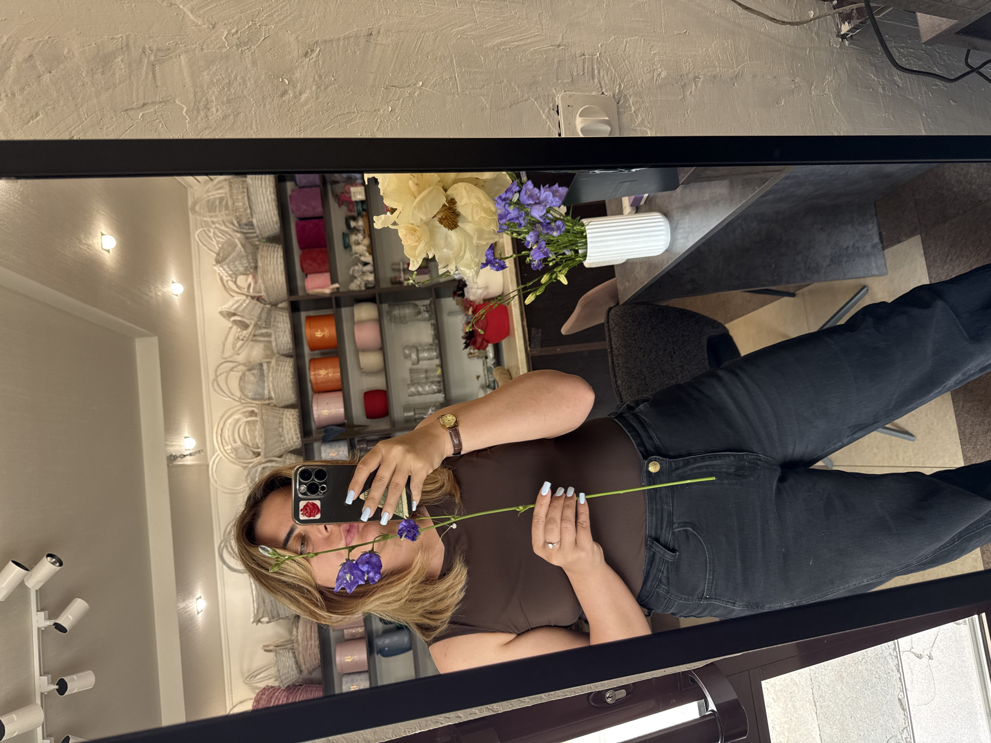 A woman takes a mirror selfie in what appears to be a flower shop. She is wearing a brown sleeveless top and black jeans. She is holding a long-stemmed flower with purple blooms in her right hand, bringing it up to her face. Her left hand is resting on her hip and she is also holding her smartphone, which is angled to capture the reflection. In the background, shelves are stocked with various baskets, vases, and decorative items. A bouquet of light-colored flowers sits on a table in front of the shelves. The floor appears to be tiled with a wood-grain pattern. The wall to the right of the mirror has a textured finish.