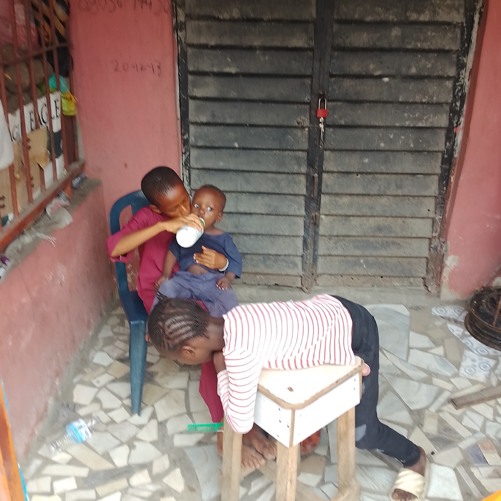The image depicts an outdoor scene in Ado, Nigeria. Three children are present. The child on the left, wearing a maroon shirt, is seated in a blue plastic chair and feeding a smaller child with a bottle. The smaller child, wearing a navy blue shirt and grey pants, sits on the lap of the child in the maroon shirt, looking towards the bottle. The third child, wearing a striped long-sleeved shirt and dark pants, is positioned in front of a small wooden table, with their head bent over and leaning on the table. The background features a closed, dark-colored shuttered door with a red padlock and key. The wall is painted pink, and the ground is covered with light-colored, irregularly shaped tiles. A discarded plastic bottle and a green comb are visible on the tiled ground. Faint red markings are present on the pink wall, with one appearing to be a date: "20-12-13". Another inscription, barely visible, seems to be "08056 774 20". The overall environment suggests an urban residential setting.