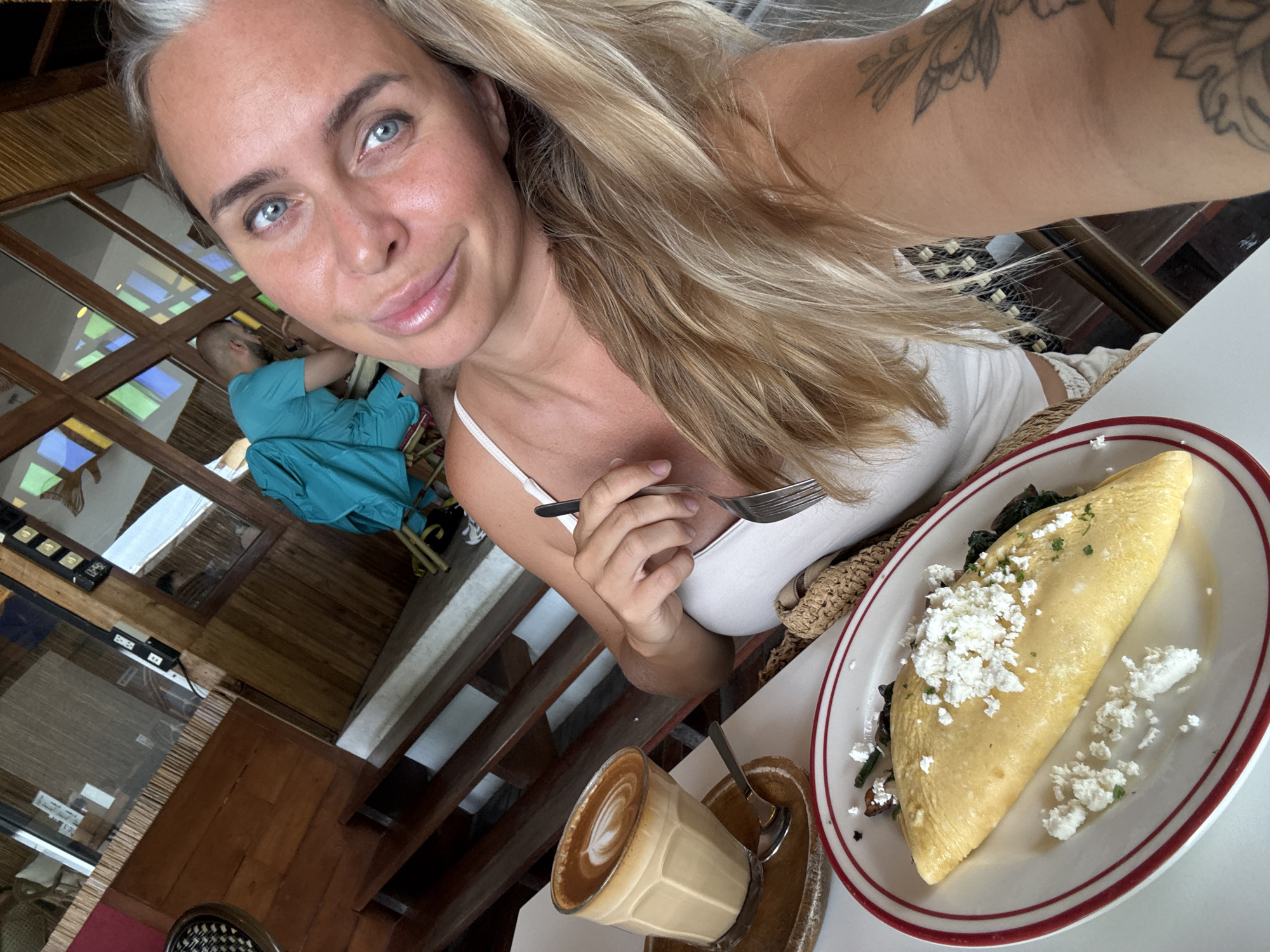 An outdoor selfie taken at a cafe in Koh Samui District, Thailand, shows a woman with blonde hair and blue eyes looking at the camera. She is wearing a white tank top. In front of her is a plate of food, possibly an omelet with cheese and greens, and a glass of coffee with latte art. The cafe has wooden furniture and bamboo accents. In the background, another person is visible, wearing a turquoise shirt. The overall setting appears to be a casual dining establishment.