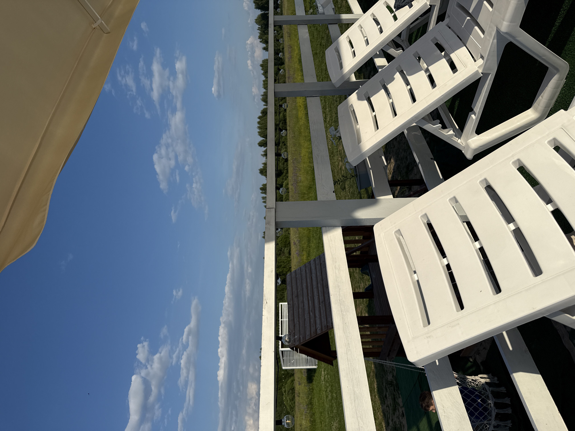 The image shows a clear blue sky with scattered white clouds, with a section of a beige umbrella in the upper right corner. Below the sky, there is a white wooden fence, and beyond it, a green field with trees in the distance. In the foreground, there are several white plastic lounge chairs. To the left of the lounge chairs, a small wooden structure with a dark roof is visible, resembling a playhouse or gazebo. A young boy is partially visible behind the fence, sitting in a white netted swing. The scene appears to be an outdoor recreational area, possibly a resort or a park, located in or near Koloksha, Russia, given the context of the prompt. The lighting suggests it is daytime, likely late afternoon or early evening due to the long shadows.