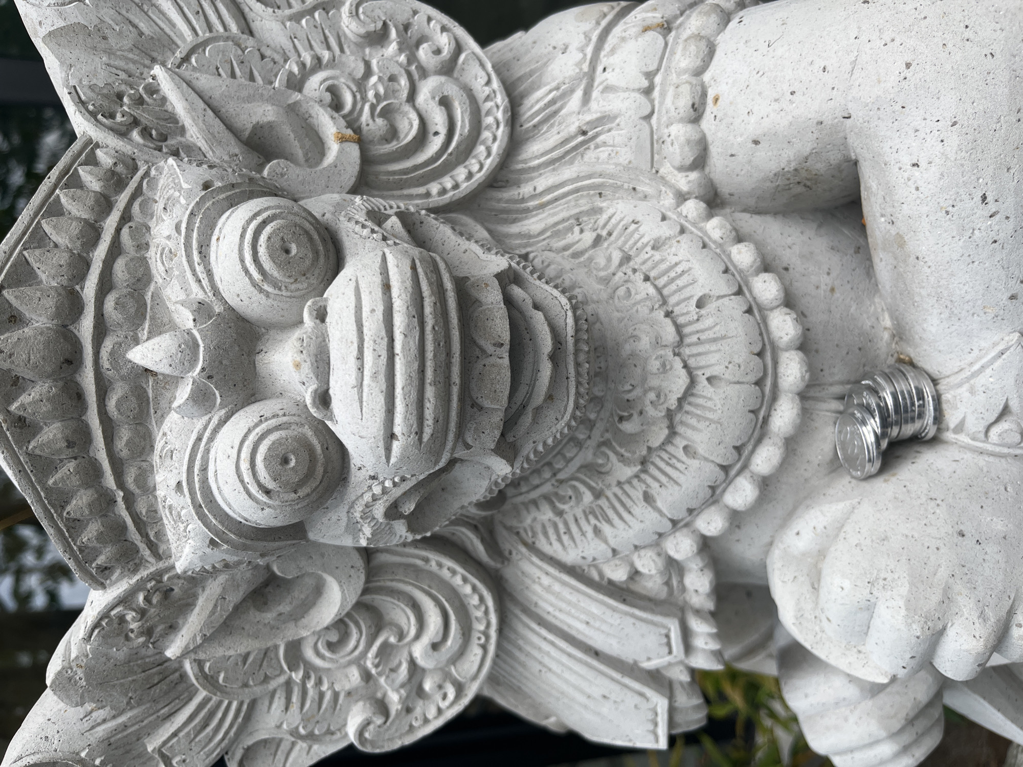 A close-up, eye-level, medium shot captures a grey stone statue of a mythological creature, likely a guardian figure, in Kuta Selatan, Indonesia. The statue is intricately carved, featuring large, bulging eyes, a wide mouth with fangs, elaborate headdresses, and feathered wings. The statue's texture is rough and porous, indicative of stone. It is positioned outdoors, with blurred greenery visible in the background, suggesting a garden or temple setting. In the lower right quadrant of the image, a small stack of silver-colored coins rests on the statue's hand or arm. The lighting is overcast, providing soft illumination and casting minimal shadows. The overall mood is solemn and stoic, emphasizing the details of the ancient-looking sculpture.