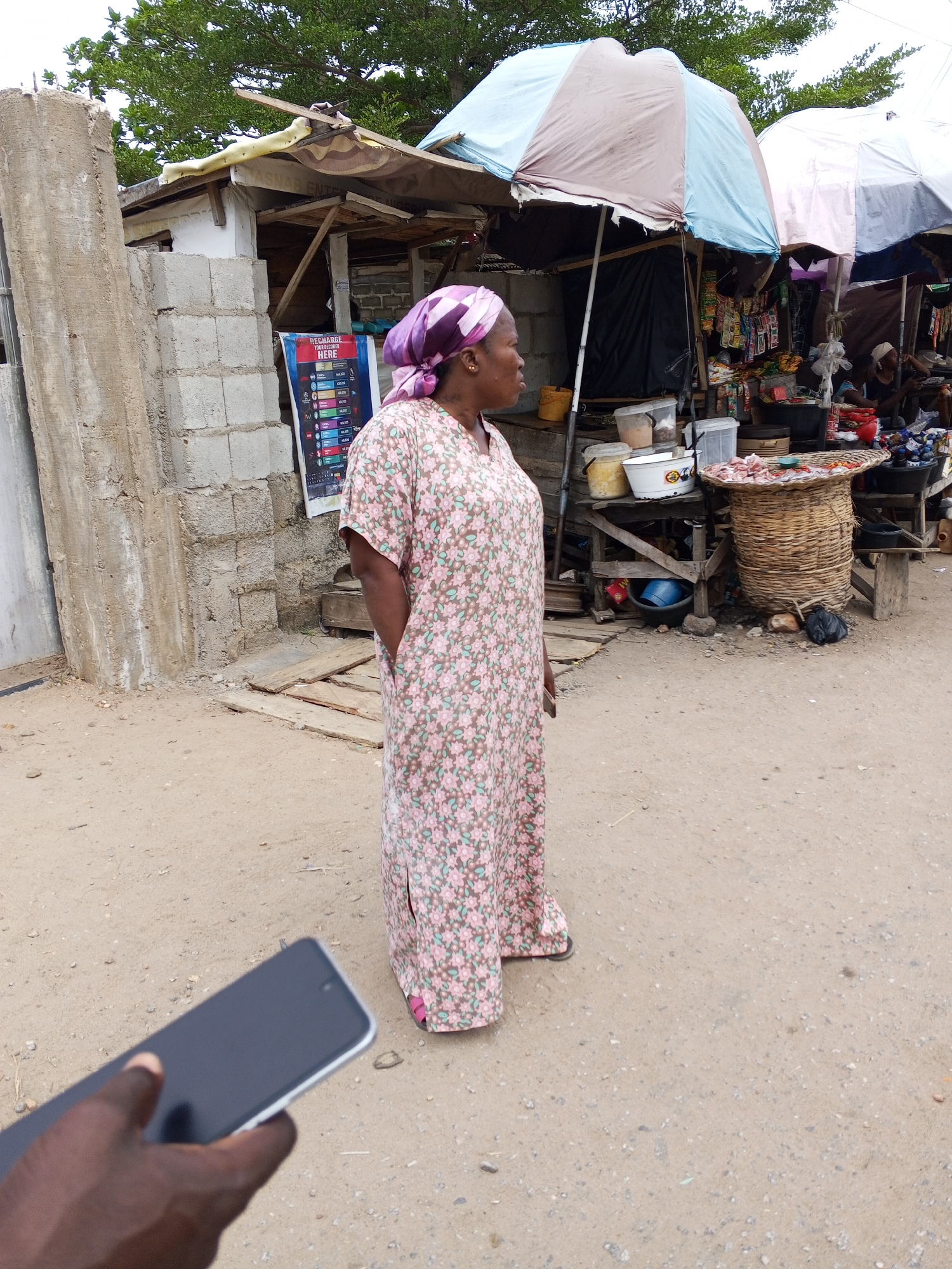 The image, captured in Zango-ndaji, Nigeria, depicts a street scene at what appears to be a local market. A woman wearing a floral print dress and a purple headscarf stands in the foreground, her gaze directed towards the right side of the frame. Her hands are placed in her pockets.
Behind her, market stalls are visible, covered by sun umbrellas of varying colors including blue, brown, and pink. A table displays various goods in containers and a woven basket overflowing with what appears to be food items. Other shoppers and merchants populate the background.
To the left, a concrete structure stands adjacent to a wooden shack. A poster is affixed to the shack.
In the lower left corner, an individual's hand holds a smartphone, partially obscuring the view. The ground is dirt and sandy.