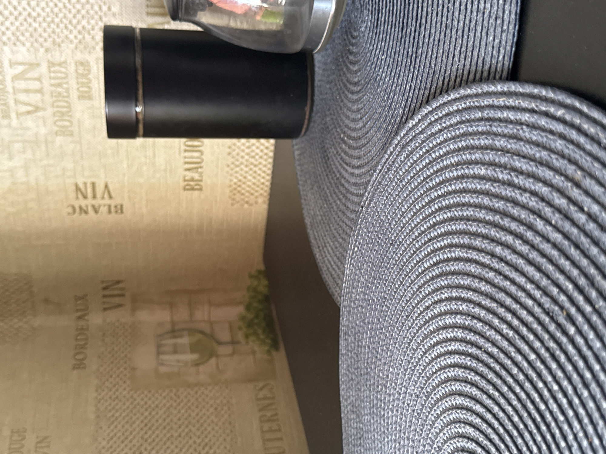 The image depicts a close-up view of two circular, gray woven placemats on a dark surface. In the background, a wall with a repeating pattern of French wine-related text and imagery is visible. The text includes words like "BORDEAUX," "VIN," and "BLANC." On the right side of the background, a tall, matte black cylindrical object, likely a container, stands next to a partially visible glass object with a metallic base. The overall scene appears to be an indoor setting, possibly a dining area. There is no indication of the specific location within Яромінскі сельскі Савет, Belarus, as the image lacks any identifiable landmarks or contextual clues.