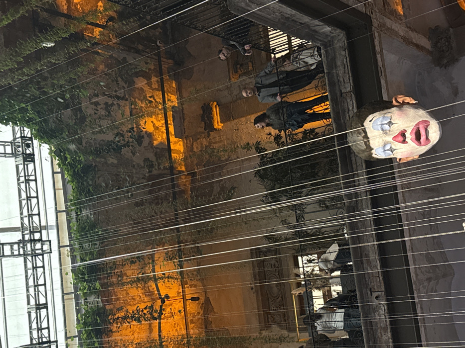 An eerie outdoor scene features a large, grotesque clown head suspended by wires. Dim light illuminates the aged building walls covered in vegetation behind it. Three individuals stand on a wrought-iron balcony above, observing the spectacle. Further down, other people are visible through another opening, creating a multi-layered perspective. The composition is slightly askew, enhancing the unsettling atmosphere.
