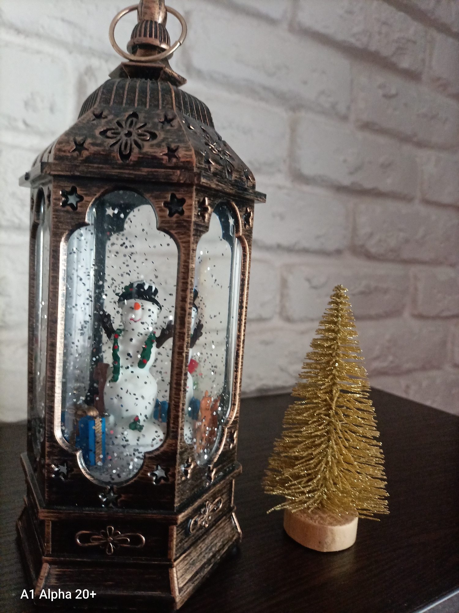 A close-up, eye-level shot shows a decorative, old-fashioned bronze lantern with a snowman inside. The lantern has glass panels with a starry design around the top. Inside the lantern, a small snowman wears a black hat, scarf, and green suspenders. Glitter is visible suspended in liquid within the lantern, suggesting it is a snow globe. To the right of the lantern, a small, golden, glittery artificial Christmas tree stands on a wooden base. The background is a white brick wall. The scene is set on a dark, reflective surface. The image is tagged "A1 Alpha 20+".