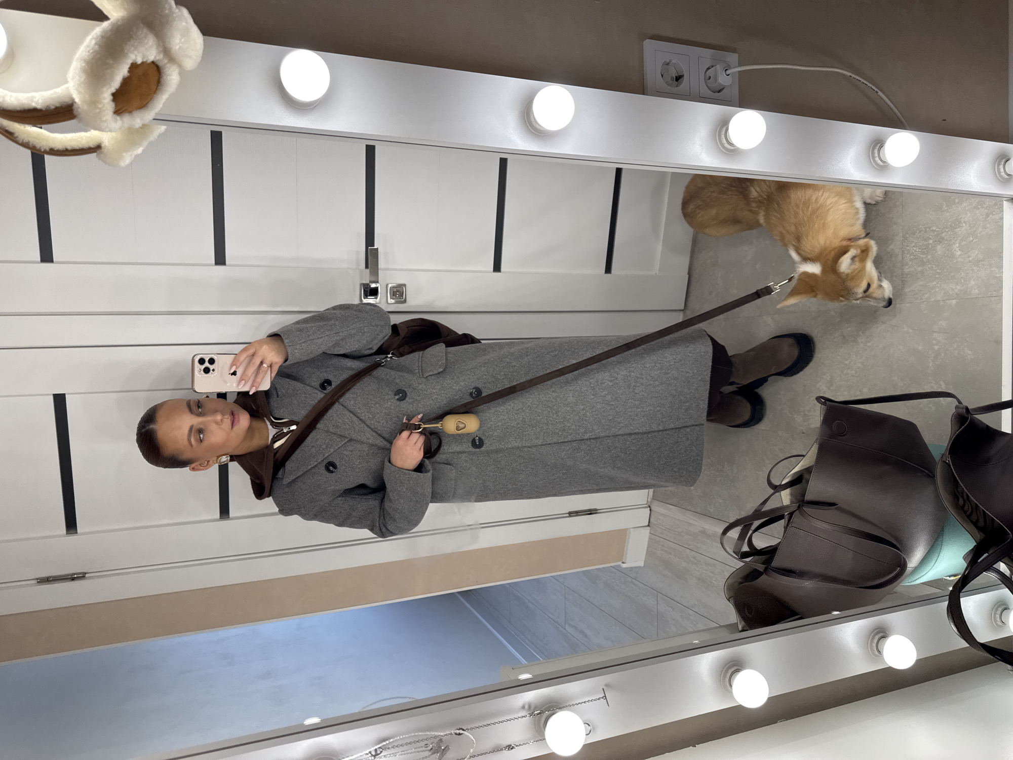 An indoor scene located in Chaikovsky, Russia, is captured through a full-length mirror selfie. A female subject, with light brown hair pulled back, wears a long grey double-breasted coat over a brown hooded top and white shirt, along with brown boots and a brown cross-body bag. She holds a pink-cased iPhone in her right hand, positioned to take the photograph, and a dog leash with an attached light brown waste bag dispenser in her left hand. A fawn and white Pembroke Welsh Corgi stands on the light-colored floor in front of the mirror, looking downwards and to the left. The white-framed mirror is illuminated by vertical rows of circular LED bulbs on both sides. Reflections reveal a white door with two black horizontal accent lines and a silver handle, a light-colored hallway beyond an open doorway to the left, and a dark brown leather tote bag resting on a turquoise surface at the bottom left. Cream-colored earmuffs with brown accents hang from the top right edge of the mirror frame. Silver necklaces are visible on a display rack reflected to the left of the mirror. A white double electrical outlet with a white power cord plugged in is visible on the right wall.