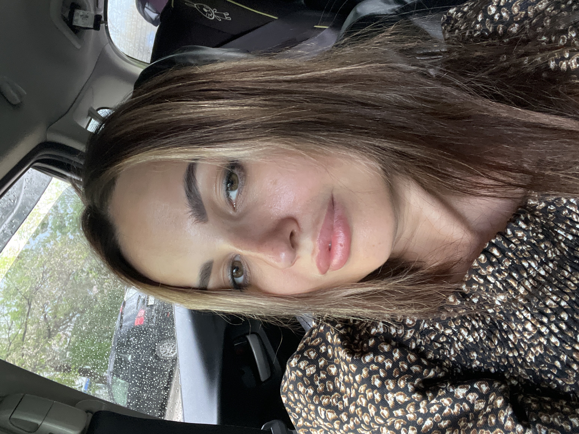 A woman with light brown hair with blonde highlights and brown eyes is pictured from the chest up, inside of a car. She is wearing a black and brown leopard print top. Raindrops are visible on the car window. A black car is visible through the window. The interior of the car is visible in the background, including a child seat.