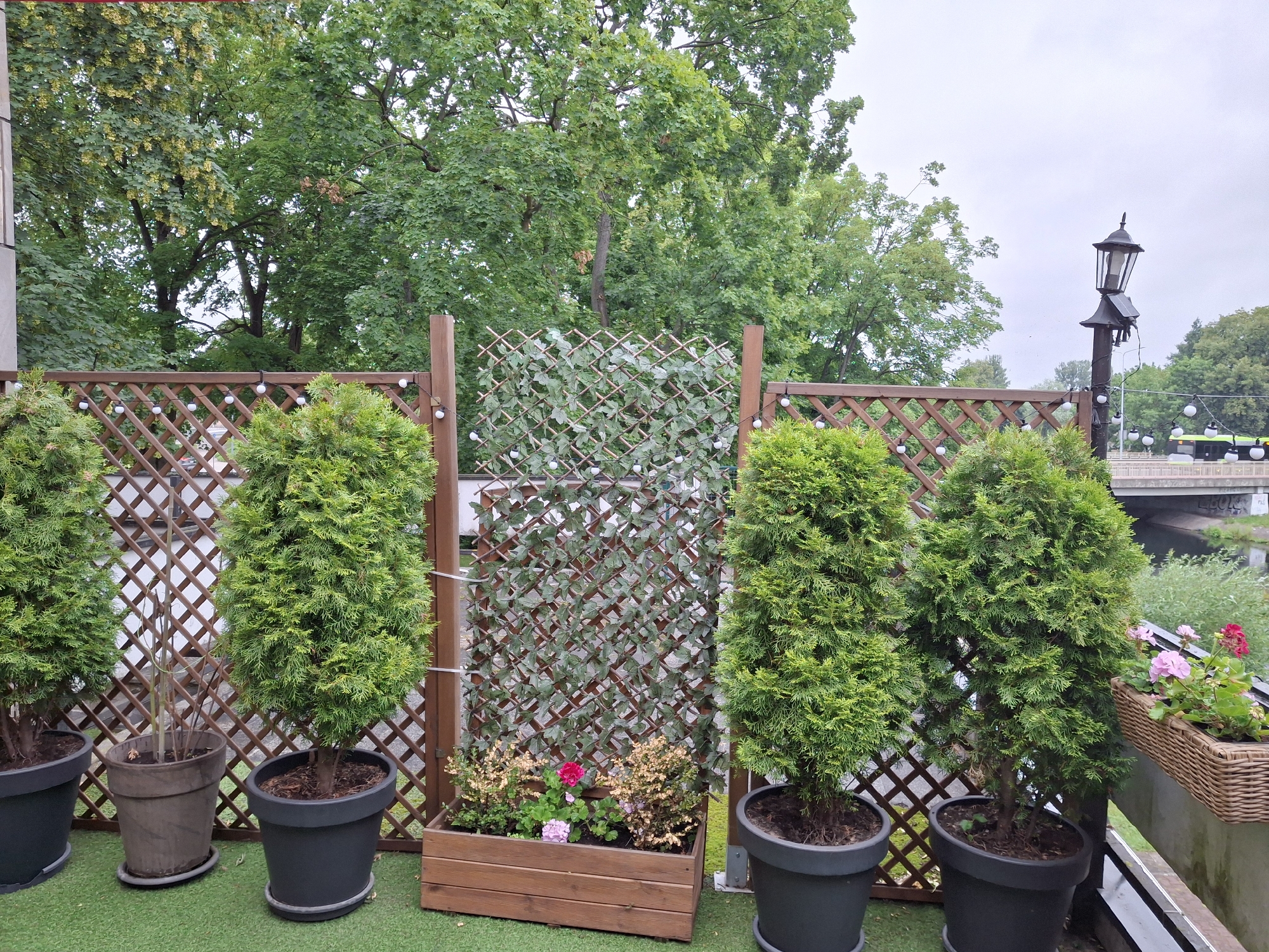 The image depicts an outdoor seating area, likely a balcony or patio, overlooking a body of water and trees. In the foreground, several potted evergreen trees are arranged in front of a wooden lattice fence. One section of the fence is covered with a green leaf-patterned screen. A wooden planter box filled with small plants and pink and red flowers sits in front of the screen. The scene is set against a backdrop of lush green trees. In the distance, a street lamp and a bridge are visible, along with a passing bus. The sky is overcast, suggesting a cloudy or possibly rainy day. There is no visible text in the image. The overall impression is one of a peaceful, natural setting with a touch of urban elements in the background.