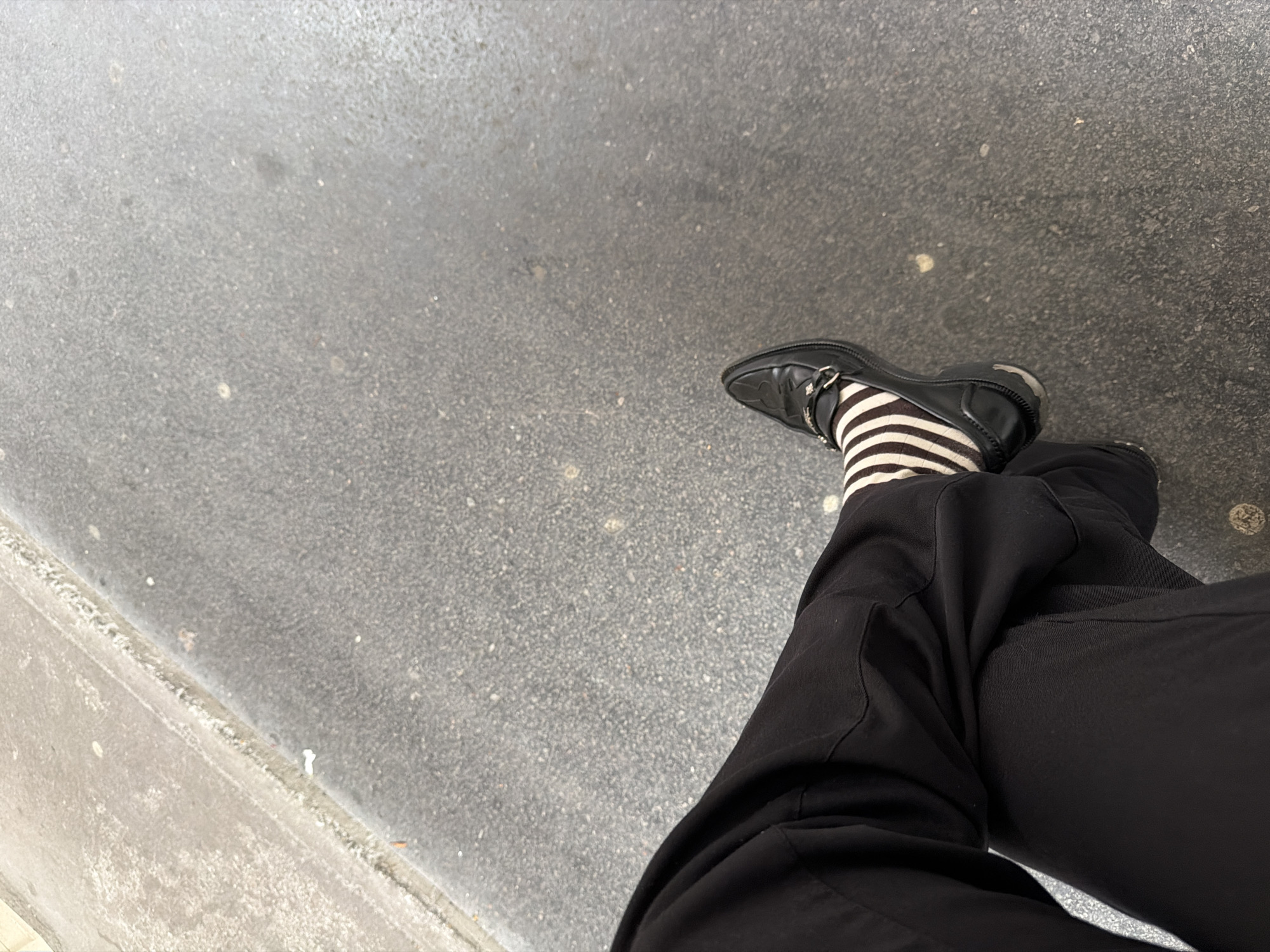 The image shows a person's legs crossed, seated outdoors on a gray concrete surface, possibly a sidewalk. The person is wearing black pants, black leather shoes with a silver buckle, and black and white striped socks. The scene appears to be in an urban environment, indicated by the concrete. While it's not possible to confirm the location as Paris, France, based solely on this image, the urban setting and typical Parisian sidewalk material make it a plausible scenario. No other people or actions are visible in the frame.