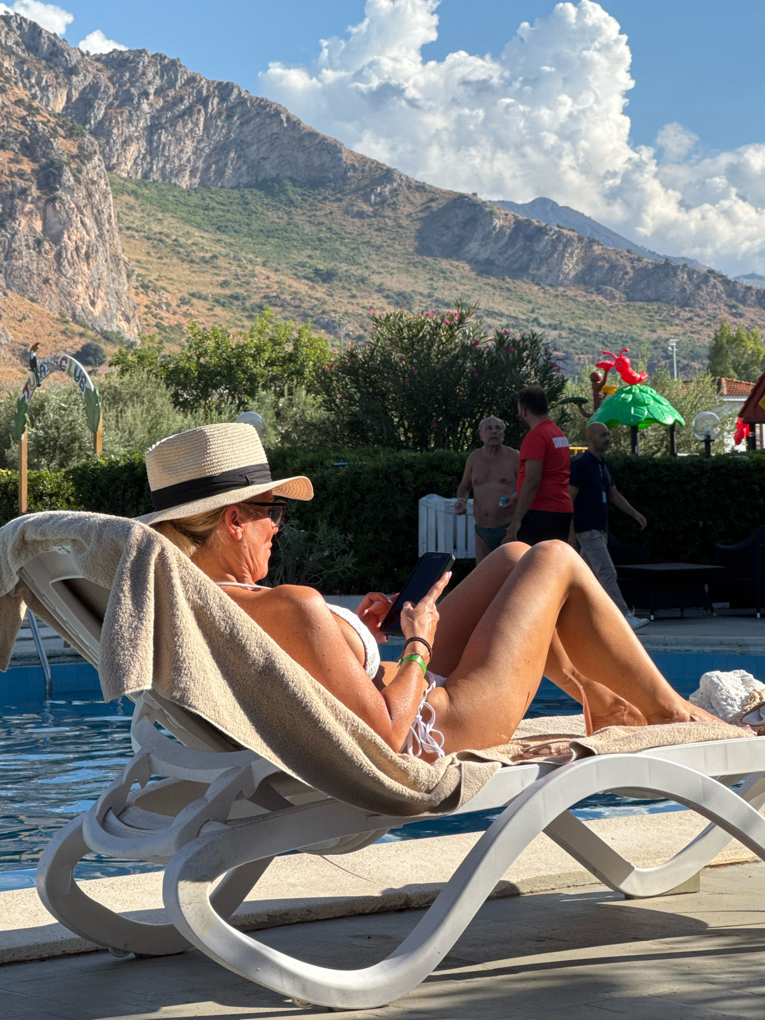 In Isola delle Femmine, Italy, a blonde woman wearing a white bikini and a straw hat with a black ribbon reclines on a white chaise lounge by a pool, holding a smartphone. A beige towel is draped over the chaise lounge. In the background, several people are visible, including a man in a red t-shirt and another man in a blue shirt. Behind them, lush green foliage and trees are set against a backdrop of rugged, rocky mountains under a bright blue sky with large, fluffy white clouds. Red and green playground structures are also present. The scene appears to be a resort or hotel area.