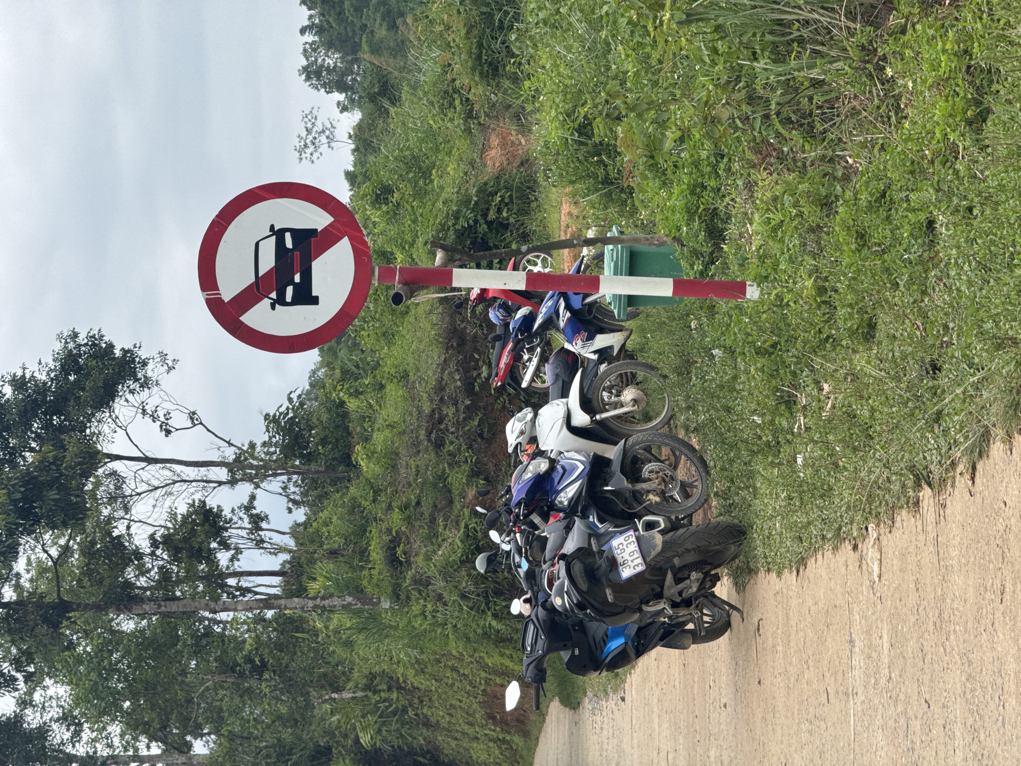 In Pù Luông Commune, Vietnam, a no-cars sign is visible on the side of a road. The sign is circular with a red border and a white background, featuring a black silhouette of a car with a red diagonal line through it. The sign is mounted on a red and white striped pole. In front of the sign, several motorcycles are parked on the side of a concrete pathway. The pathway is bordered by lush green vegetation and trees. The sky is overcast.