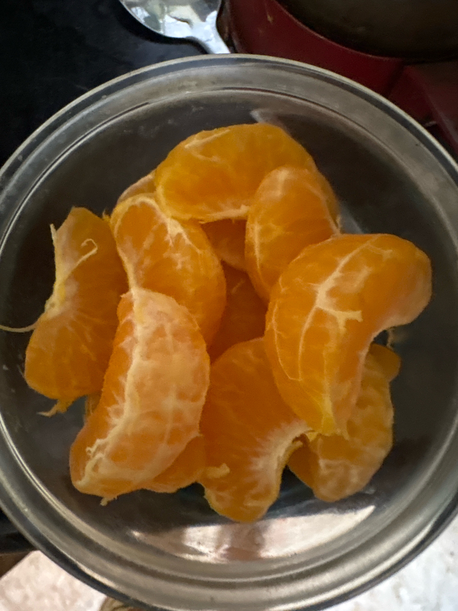 A close-up, overhead shot shows peeled orange segments arranged in a clear glass bowl. The segments are bright orange and appear juicy. The bowl is placed on a dark, textured surface, with hints of a red appliance in the upper right corner and a metallic spoon in the upper left. The scene is dimly lit, with soft light illuminating the fruit. There is no indication of the city of Ashvem, India.