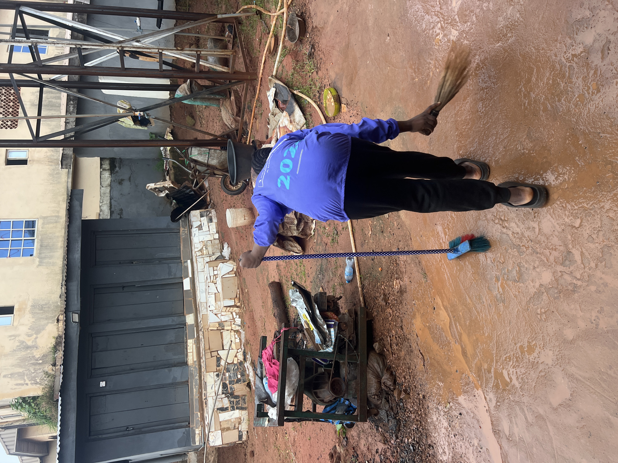 The image depicts a scene in Nkpor, Nigeria, with a person in a purple jacket and black pants sweeping a muddy ground. The person is holding a broom with a blue and white dotted handle. In the background, there is a building with dark doors, a generator on a metal stand, and a metal water tower. The ground is covered in red mud and puddles of water, suggesting recent rain or poor drainage. Debris and various objects, including a plastic bottle and a tarp, are scattered around the area. The overall environment appears to be an outdoor space adjacent to buildings, possibly a workshop or a commercial area.