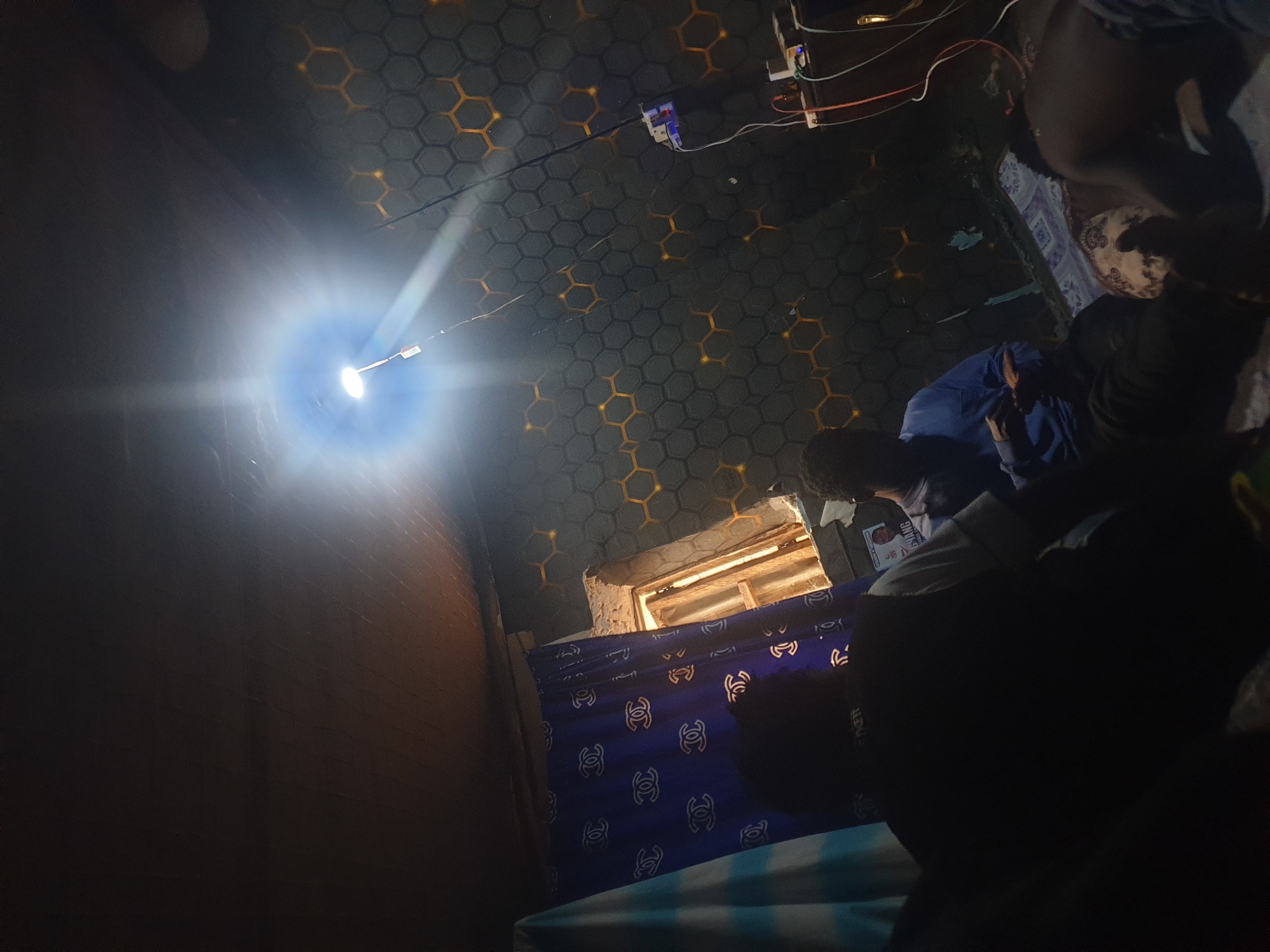The image shows an indoor scene, dimly lit by a bright overhead light source. The walls are decorated with a hexagonal pattern. To the left, a blue curtain with a repeating pattern of "C" logos is visible. A small window with wooden frames is present. In the foreground, three individuals are seated or lying down. One person in the center wears a blue shirt and appears to be looking at a device in their hands. Another person is seen from the back, wearing a black shirt with lettering on the collar. A third individual is partially visible in the lower right corner. Electrical outlets and cords are mounted on the wall on the right side of the image. The setting does not provide specific indicators of the city of Washington, United States of America.