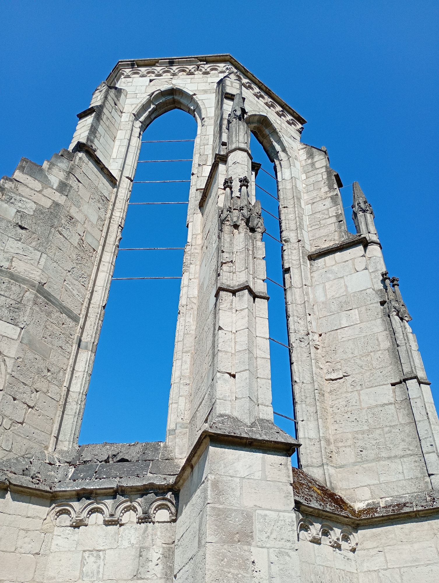 The image shows a low-angle view of the ruins of a large stone structure, likely a church or abbey, against a clear blue sky. The stone is a light, weathered gray and is constructed in large blocks, exhibiting architectural features characteristic of Gothic style, such as pointed arches and decorative stonework. The prominent feature is a massive, open window frame, revealing the sky. The structure appears damaged and incomplete, with sections missing and the stonework showing signs of age and decay. Metal bars have been installed across the opening of the window, likely for structural support or to prevent further collapse. There are no people or animals visible in the image. The scene is static and conveys a sense of historical preservation and architectural grandeur. This ruin is part of the Walkenried Abbey complex located in Walkenried, Germany.