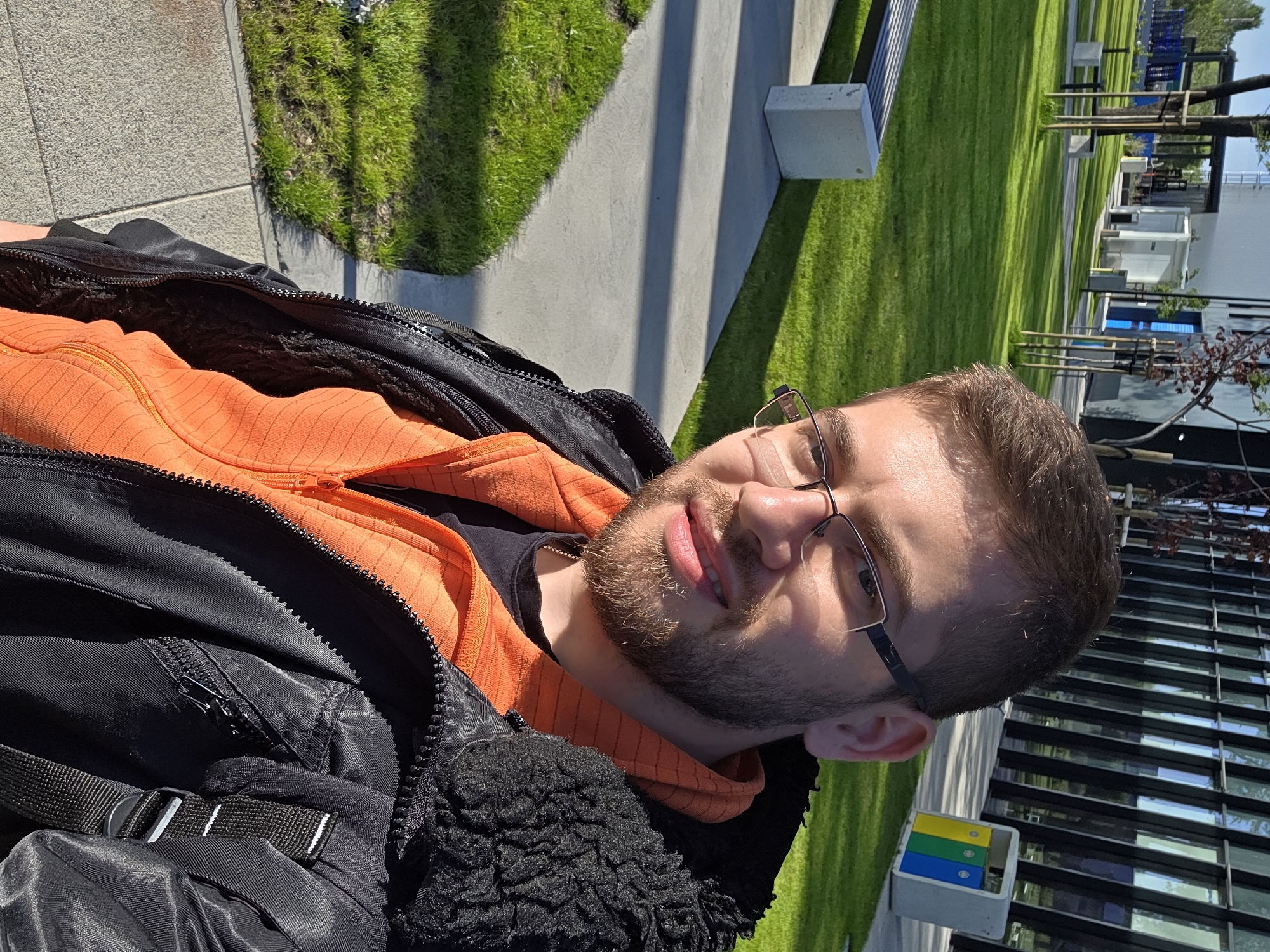 A young man with light brown hair, a short beard, and glasses is smiling gently at the camera. He is wearing a bright orange hooded sweatshirt with a subtle striped texture, layered under an open black jacket that features prominent zippers and a black furry trim around the collar or opening. A black strap with a white-striped buckle is visible on his left shoulder.
The scene takes place outdoors on a sunny day, likely in a modern public park or university campus setting. The background features well-maintained green lawns, intersected by smooth concrete pathways. Long shadows are cast across the grass and paths, indicating strong sunlight, possibly in the morning or late afternoon. Several contemporary benches, some made of concrete blocks and others with slatted designs, are scattered throughout the grassy areas. In the distance, a multi-story building with a facade composed of numerous windows reflects the bright light. Small trees with bare branches are visible, suggesting either early spring or a type of tree with sparse foliage. Towards the bottom right, a grey recycling bin with three distinct color-coded compartments (yellow, blue, green) stands near the building.
The man appears relaxed and content, posing for the photograph. The overall atmosphere is bright and pleasant. While the exact location is Plovdiv, Bulgaria, no specific landmarks confirming this are visible in the image itself.
