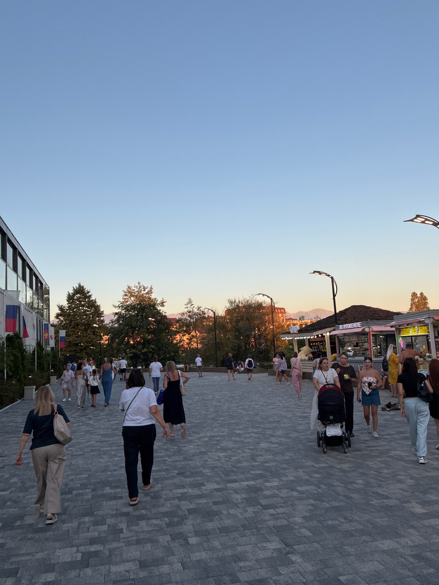 The image depicts a scene on a paved pedestrian walkway in Sochi, Russia, likely during the evening or late afternoon, given the clear sky and soft lighting. The walkway is made of large, grey, rectangular pavers. On the left side of the image, a modern building with a glass facade and dark window frames is visible, adorned with flags of Russia and another country, possibly the United Kingdom or France. Several people are walking on the pathway, some individually and others in groups. A woman in a dark top and beige pants is walking away from the camera on the left. Further down the walkway, a woman in a white top and dark pants is walking towards the camera, pushing a stroller. To the right, there are several small stalls or kiosks, one of which has Russian text above it, suggesting a food or souvenir vendor. A person in a yellow dress stands near one of the kiosks, and a shirtless man is visible behind another. People are casually dressed, indicating a relaxed atmosphere. Trees with green foliage are present in the background, and a mountain range is faintly visible on the horizon, bathed in the warm glow of the setting sun. Modern streetlights with an artistic design are also visible, illuminating the area.