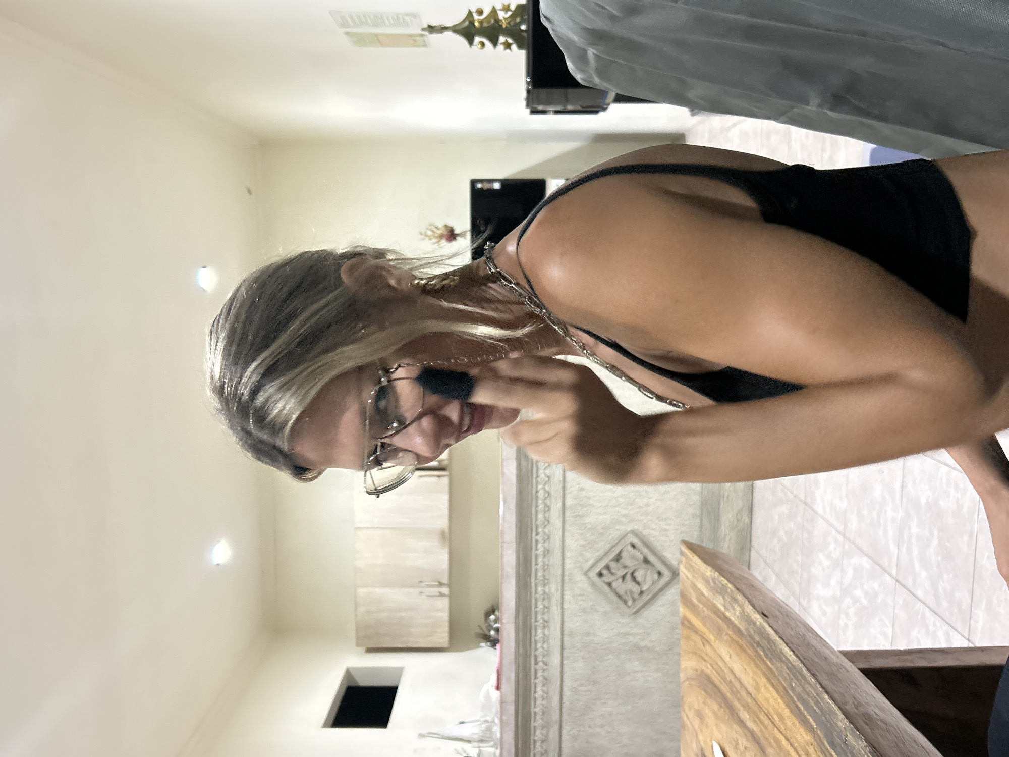 The image, correctly oriented, depicts a woman indoors, engaged in what appears to be makeup application. She has blonde-streaked hair, wears glasses with clear frames, and is dressed in a black top or swimsuit, accessorized with a silver-colored chain or necklace around her neck. She holds a dark makeup brush to her face, focusing intently.
The scene is an interior room, possibly a kitchen or dining area, with artificial lighting suggesting evening or night. The background features light-colored wooden cabinets on the left, with a dark rectangular opening below them. To the right, a decorative wall panel with an intricate carved pattern is visible. A large wooden furniture piece with a distinct grain occupies the foreground on the right. The floor appears to be white-tiled.
A reflective surface in the upper background mirrors a golden, crown-like trophy or ornament. Partially visible, reflected text on this surface spells out "JIHIL". The location is contextually identified as North Kuta, Indonesia.