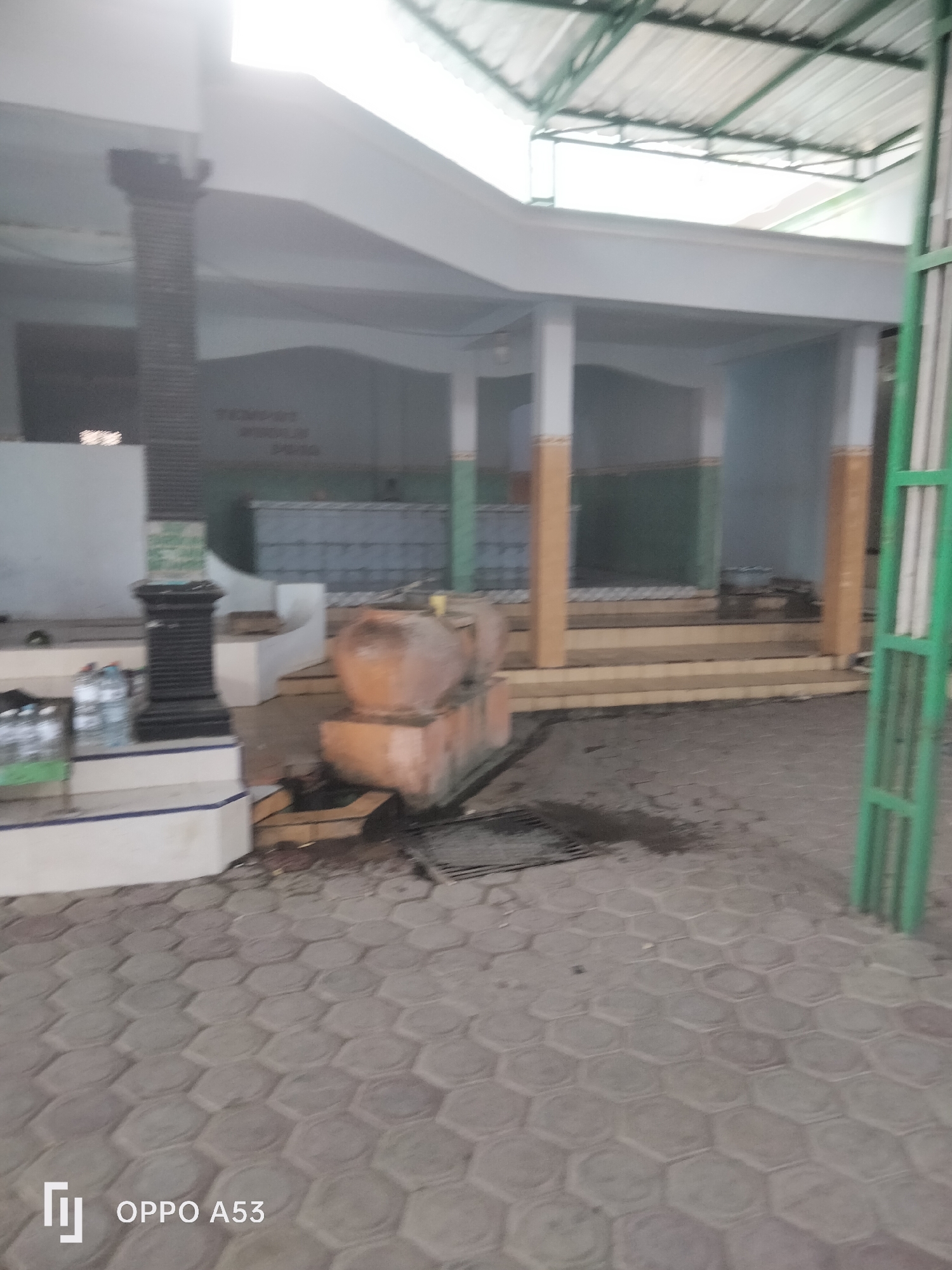 The image depicts a covered outdoor area with a tiled floor made of hexagonal pavers. In the foreground, a series of plastic bottles are visible on a raised platform. To the right, a section of a green metal fence is present. Behind the platform, a large, terracotta-colored urn or basin sits on a concrete structure. The background reveals a covered walkway with columns. Faint text on a wall in the background reads "TEMPAT WUDHU POO". The overall scene suggests a public facility, possibly a place of worship or a rest stop, located in the city of Badung, Indonesia.