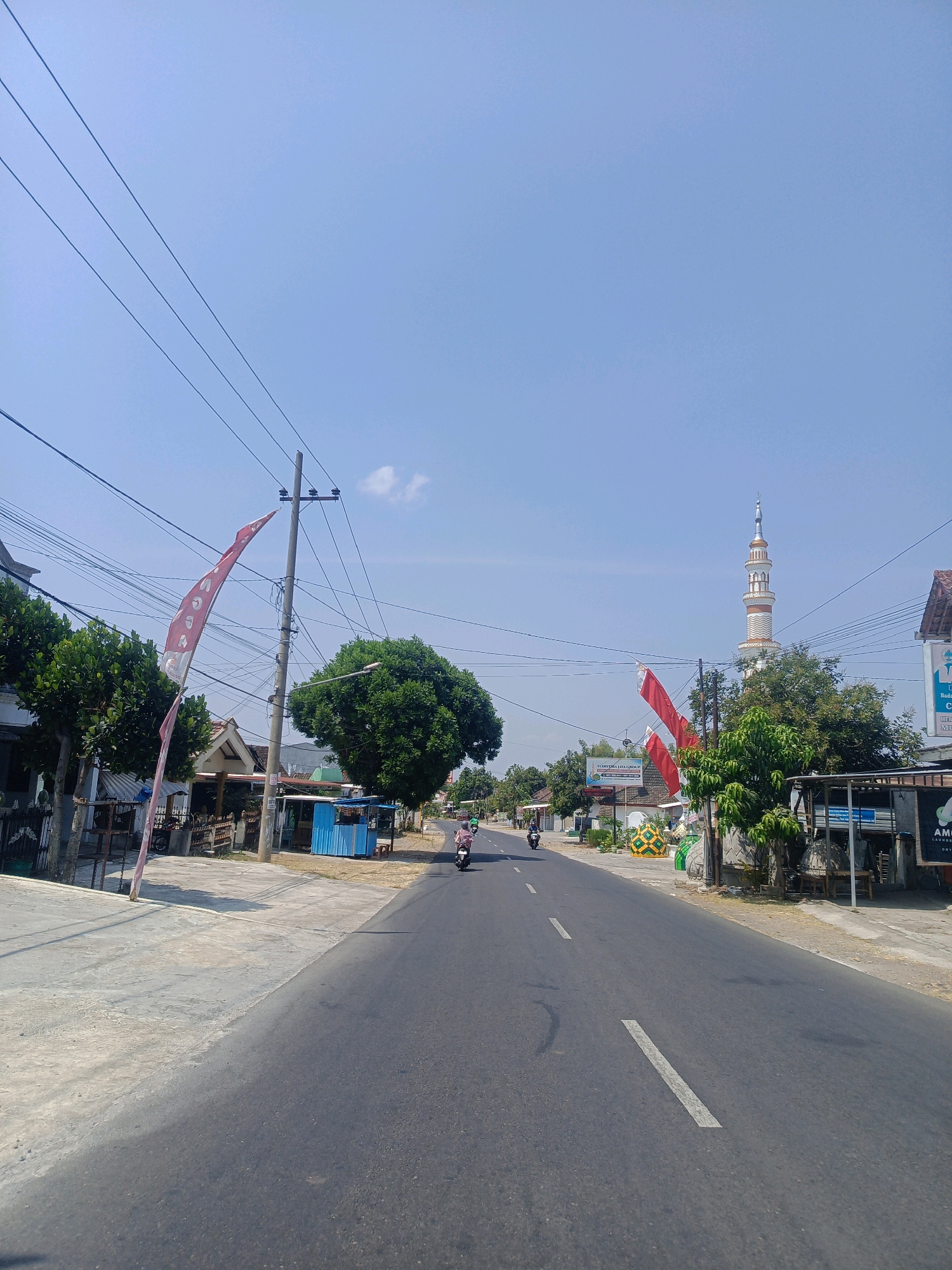 The image captures a street scene in Milangasri, Indonesia, under a clear blue sky with scattered clouds. The road, paved with asphalt, runs through the center of the frame, dividing the landscape. Along the right side of the road, a mosque with a tall, ornate minaret stands prominently. Red and white flags, possibly national flags, are displayed on poles, adding a festive touch to the environment. Various buildings, including small shops and houses, line the street, some with trees and vegetation in front of them. A few individuals are seen riding motorcycles, contributing to the sense of local activity. Power lines crisscross the sky, typical of the area. The overall impression is of a bright, sunny day in a developing urban or semi-urban setting.