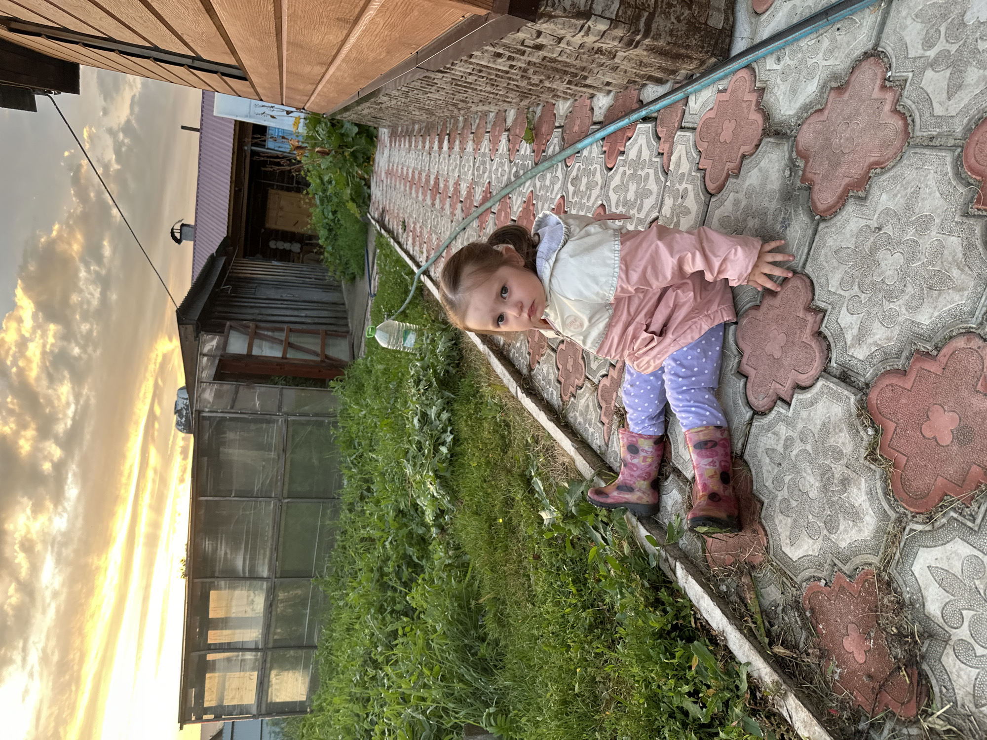 A young girl with brown hair pulled back in a ponytail is sitting on patterned paving stones outdoors. She is wearing a pink and white jacket, purple polka dot leggings, and patterned rain boots. The girl is looking at the camera with a serious expression. To her left, a hose is visible running along the ground, and a plastic water bottle lies nearby. Behind her, lush green vegetation grows, and a greenhouse structure with glass panes is visible. Further in the background, a rustic wooden building with a red-tinted metal roof stands against a sky filled with dramatic, sun-drenched clouds, suggesting it is either sunrise or sunset. The paving stones are a mix of grey and reddish-brown tiles with floral patterns. The scene appears to be in a rural or semi-rural setting, possibly a backyard or garden area in Yaroslavka, Russia.