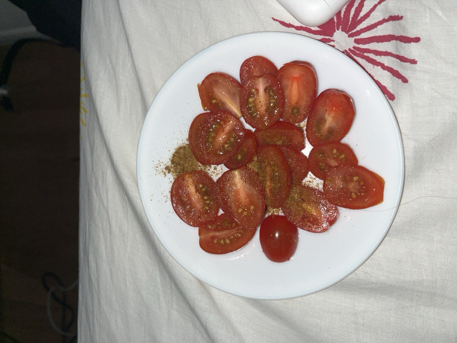 A white, circular plate holds approximately 18-20 red tomato slices and one whole red tomato, all topped with a scattering of brownish granular seasoning. The cut tomato surfaces reveal internal pulp and pale seeds. A minor linear imperfection, possibly a scratch or crack, is visible on the bottom rim of the plate. The plate rests on a light-beige woven fabric surface displaying wrinkles and folds. To the immediate right of the plate, on the fabric, a partial reddish-pink radial pattern is visible. A white, rounded rectangular object, consistent in appearance with an Apple AirPods charging case, is partially visible further to the right. The background area above the fabric is dark and indistinct, indicating a dimly lit indoor space with blurred shapes. No individuals are present within the frame. The image does not contain any discernible geographical markers or architectural features to establish a specific location within the city of Strasbourg, France; it depicts a generic indoor setting.