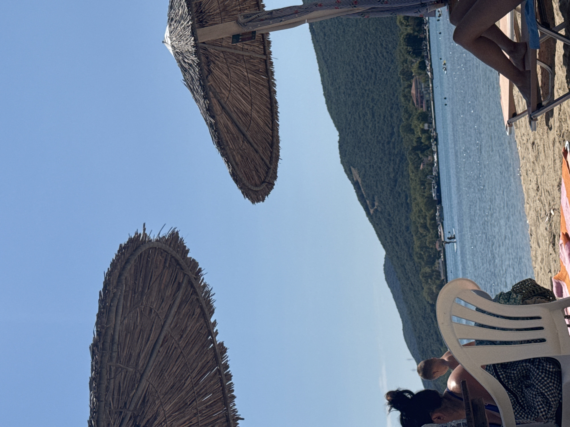 The image depicts a sunny day at a beach in Stavros, Greece, with the turquoise waters of the Aegean Sea stretching towards a verdant, tree-covered mountain range in the background. Two large, thatched beach umbrellas dominate the foreground, casting dappled shadows on the sandy shore. In the left foreground, a woman with her hair tied up in a bun sits at a table, partially obscured by a white plastic chair. A young child stands behind her, looking out towards the sea. To the right, a person's legs are visible, resting on a beach lounger beneath another umbrella. In the water, two individuals are visible on a paddleboard, and several other small boats are dotted across the surface. Buildings and more trees line the shore of the bay. The sky is a clear, vibrant blue.